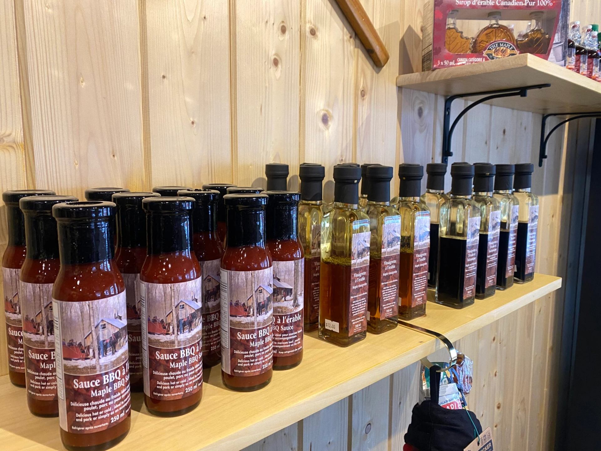 Shelves displaying bottles of maple BBQ sauce and maple vinegar inside a rustic wooden shop, with other maple products visible in the background.