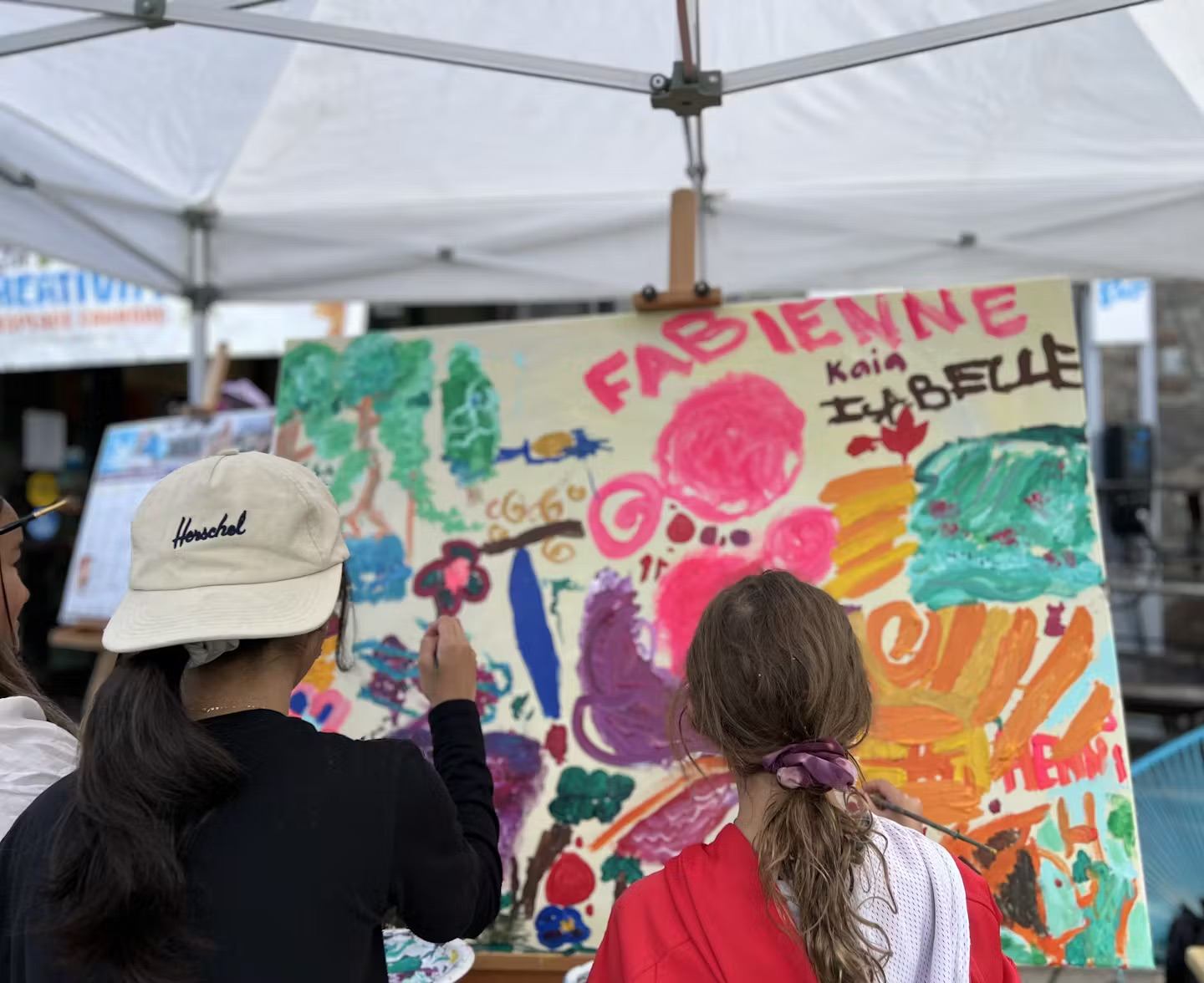 Two children paint on a colorful community art mural under a white tent at an outdoor event.