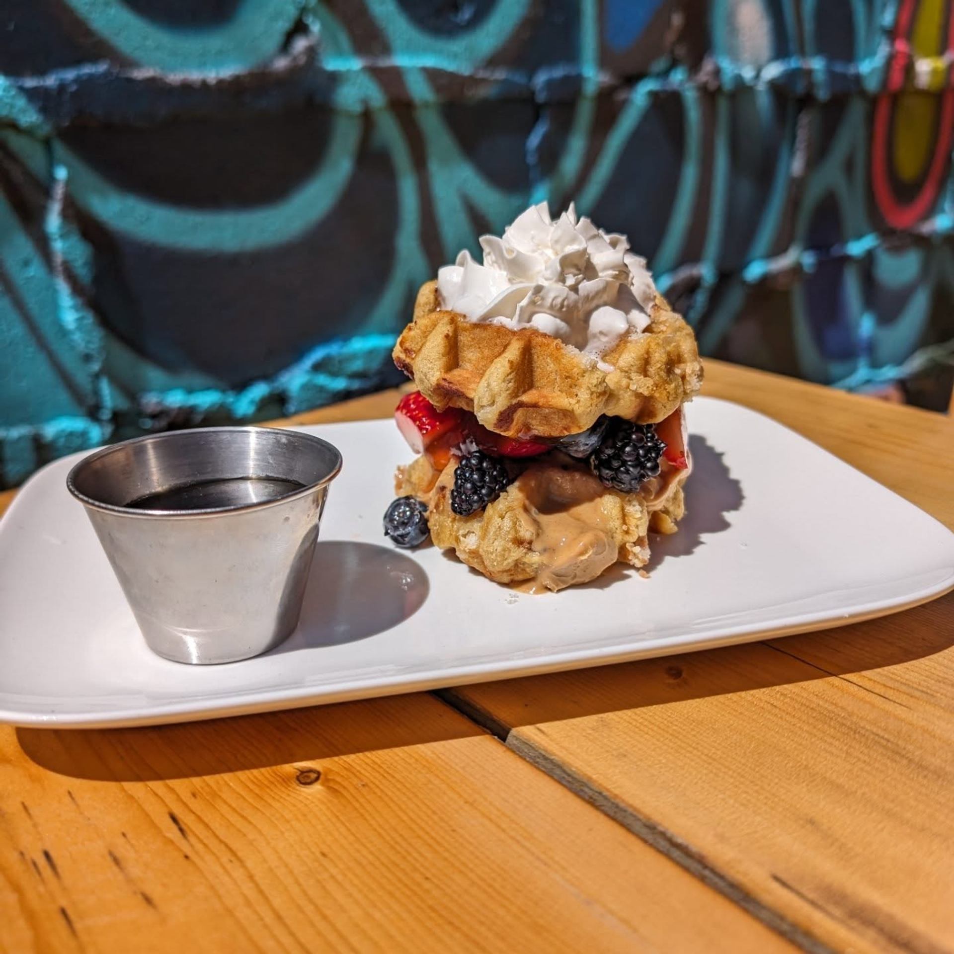 Berry waffle sandwich with whipped cream and syrup is served on a white plate atop a wooden table.