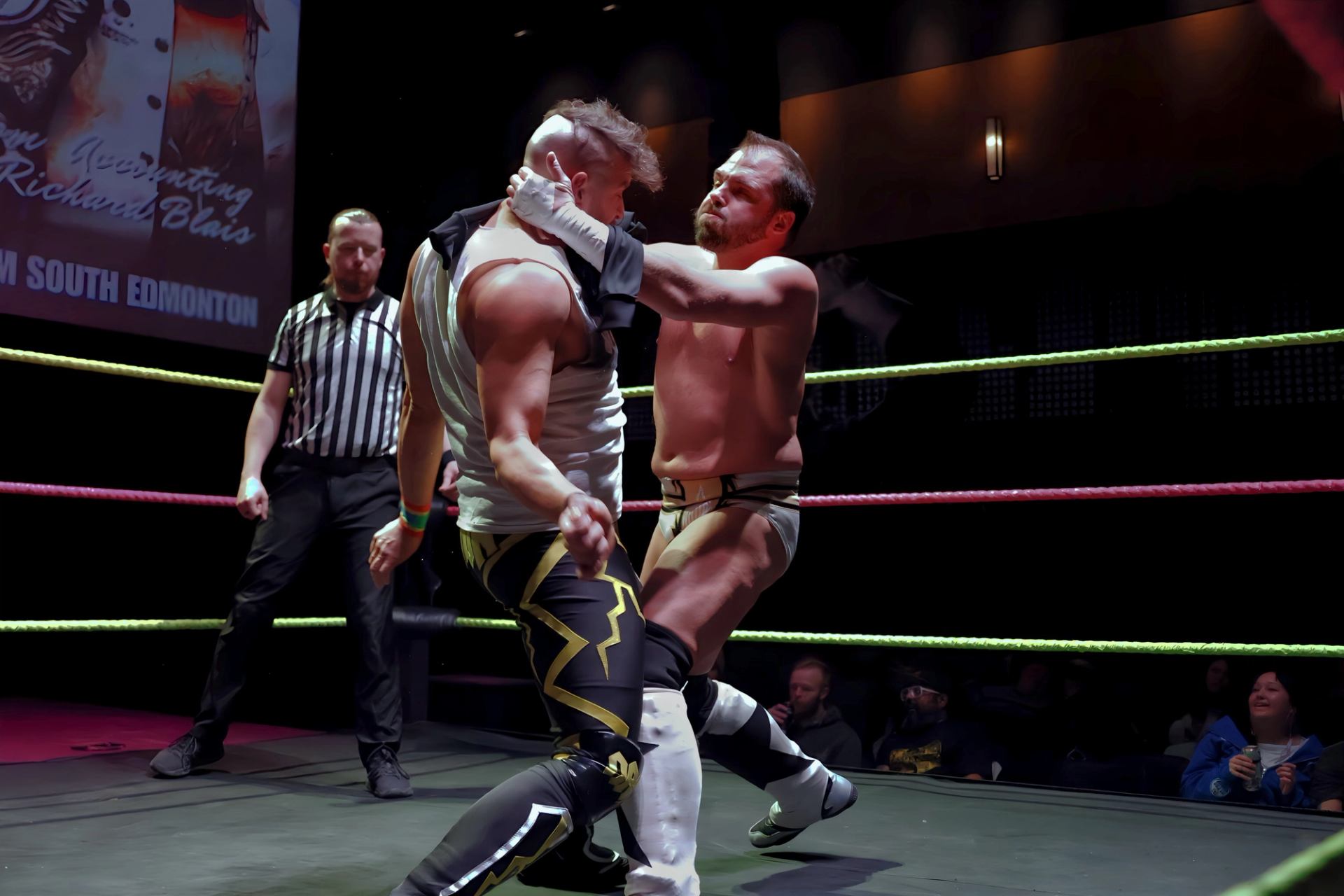 Two wrestlers grapple in ring while referee watches closely during match.