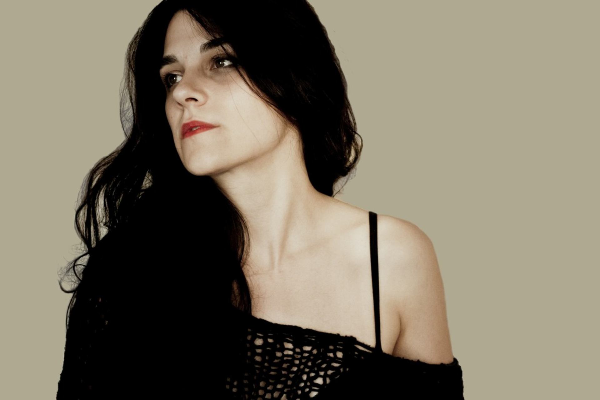 Person with long dark hair in black top looks to the side against beige background.