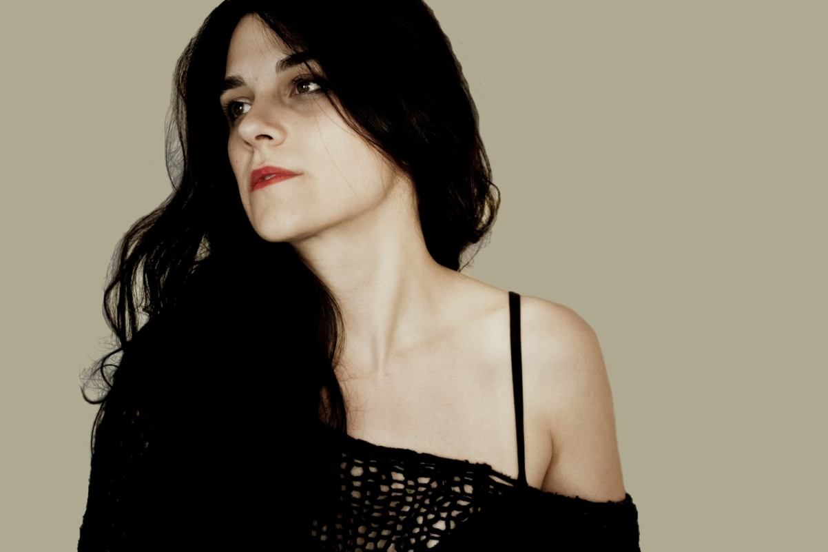 Person with long dark hair in black top looks to the side against beige background.