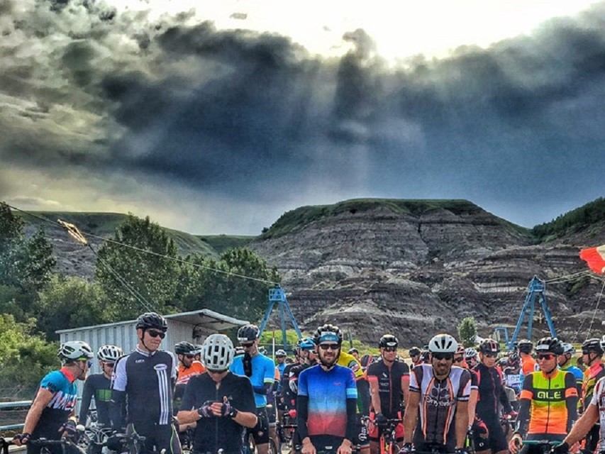 Cyclists in colourful jerseys gather with bikes before hills under dramatic cloudy sky.