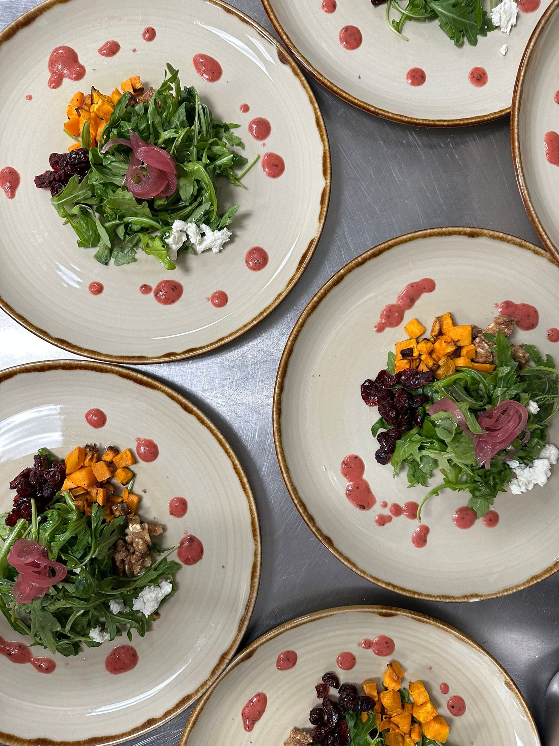 Plated arugula salads with roasted squash, goat cheese, cranberries and pink dressing dots.