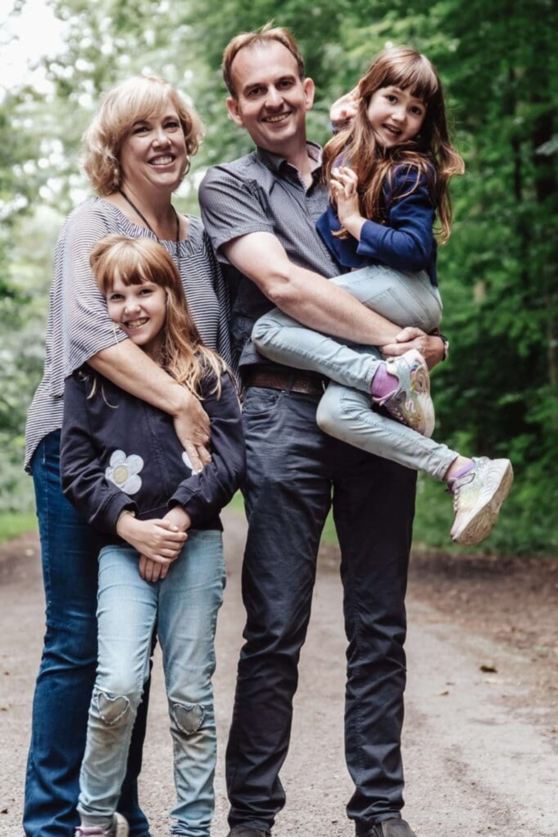 Two adults and two children stand on a forest path, one child being carried.