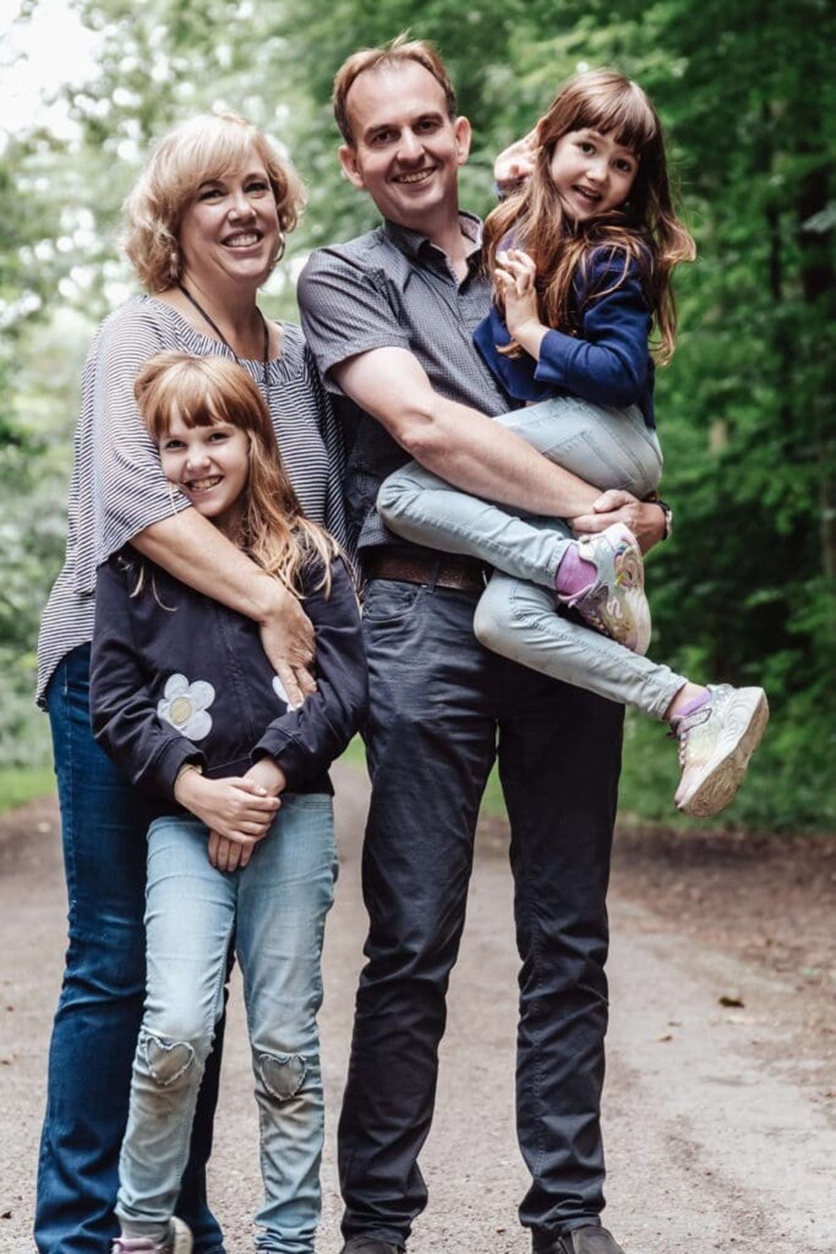 Two adults and two children stand on a forest path, one child being carried.