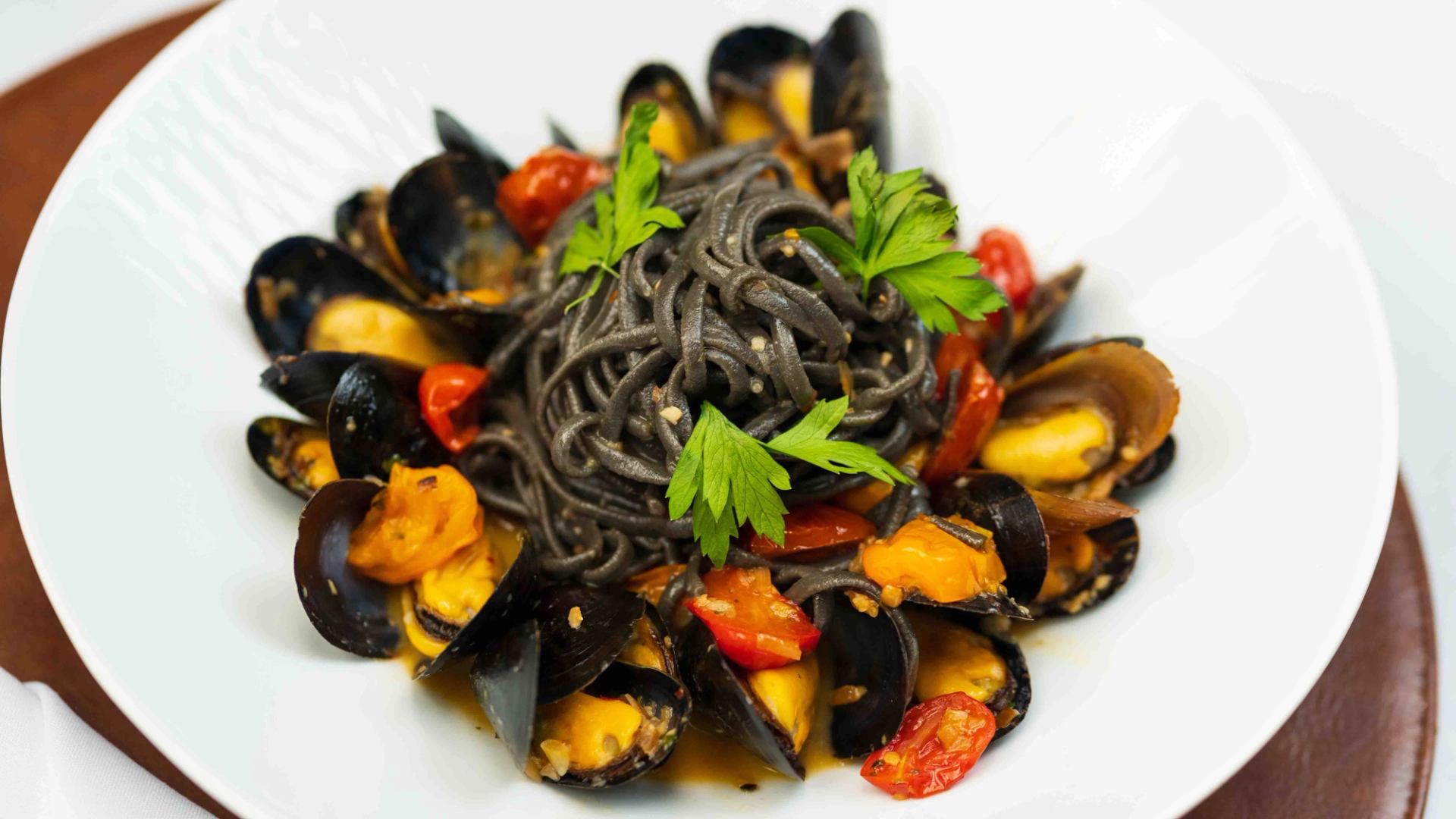 Black spaghetti with mussels, cherry tomatoes and parsley in a white bowl.