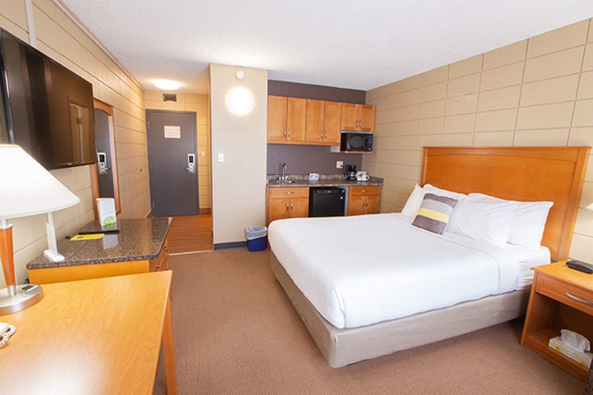 A hotel room includes a neatly made bed, a flat-screen TV, and a kitchenette with cabinets, microwave, and mini fridge. A desk and lamp sit in the foreground, emphasizing a practical setup.