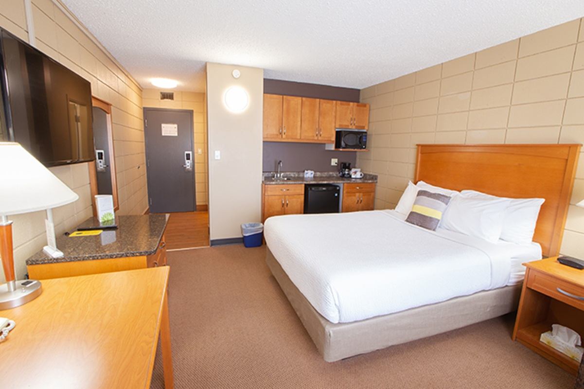 A hotel room includes a neatly made bed, a flat-screen TV, and a kitchenette with cabinets, microwave, and mini fridge. A desk and lamp sit in the foreground, emphasizing a practical setup.