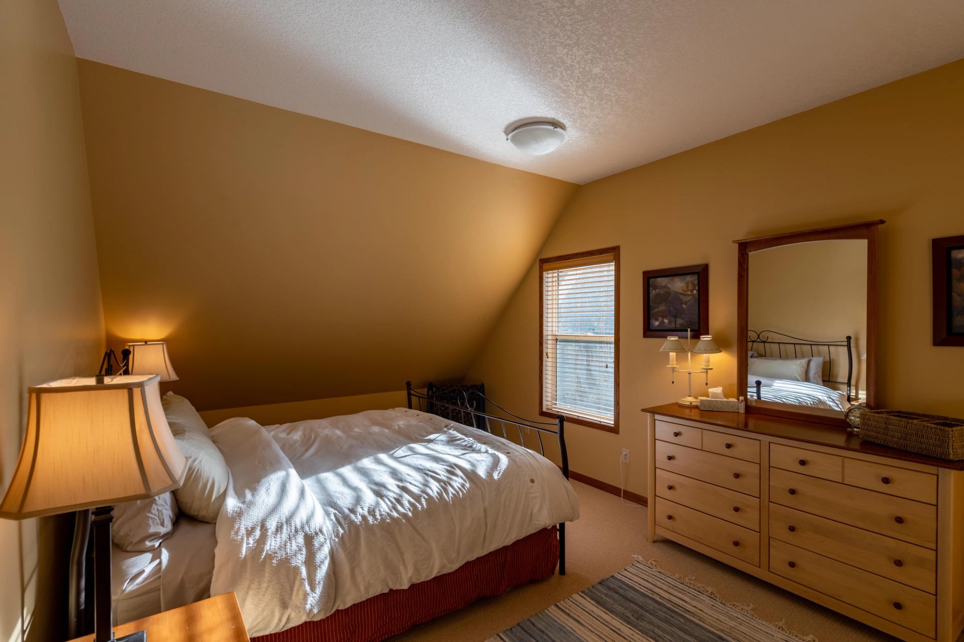 Bedroom with double bed, bedside table, dresser, and window blinds in warm neutral walls.