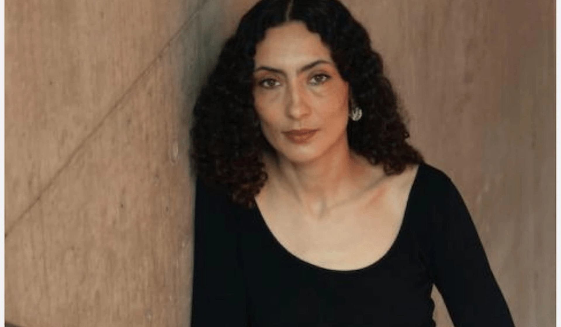 Person with curly hair in black top sits against a plain beige wall.