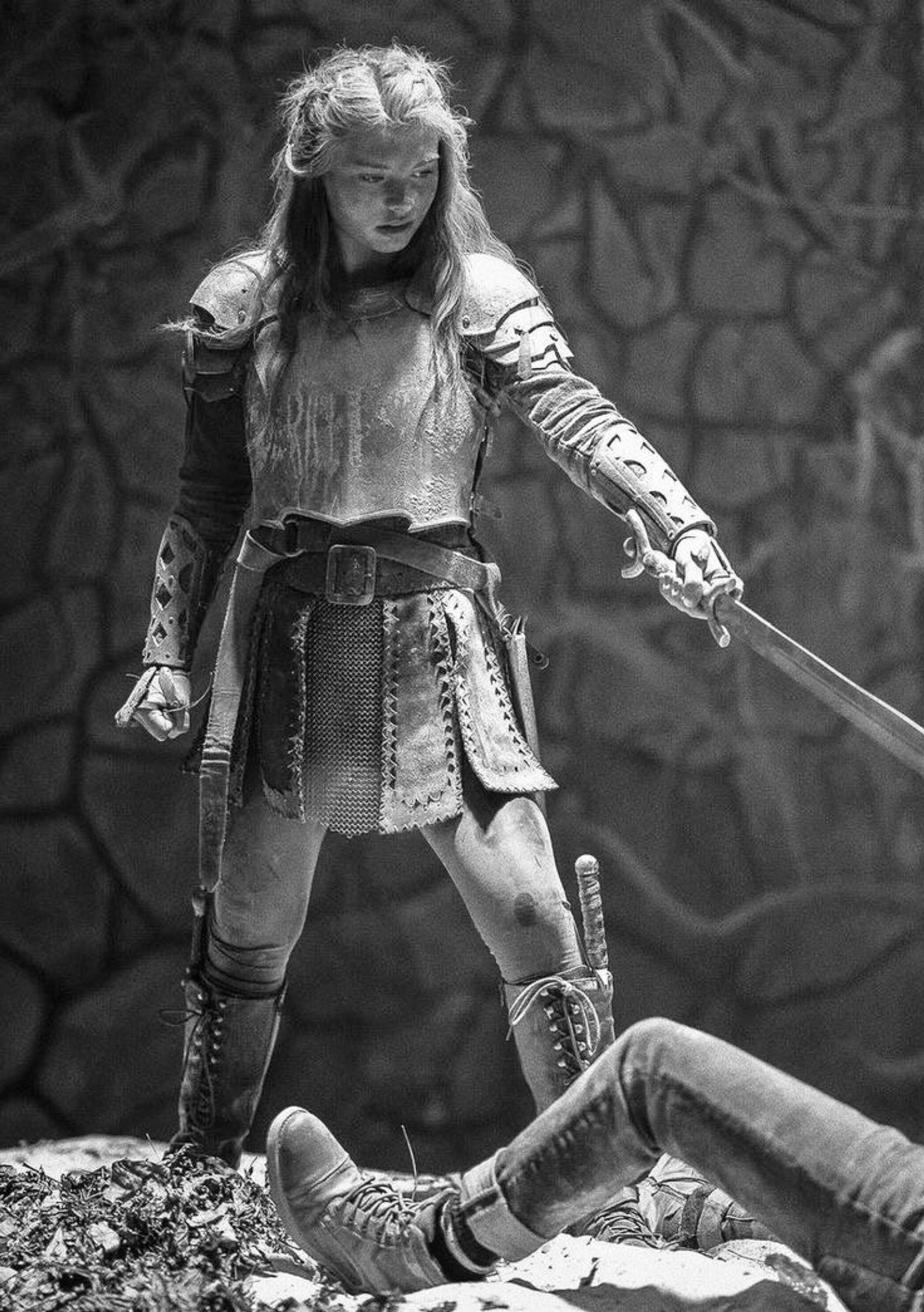 Black and white image of a young girl in armor with a sword, standing over a fallen person's legs.