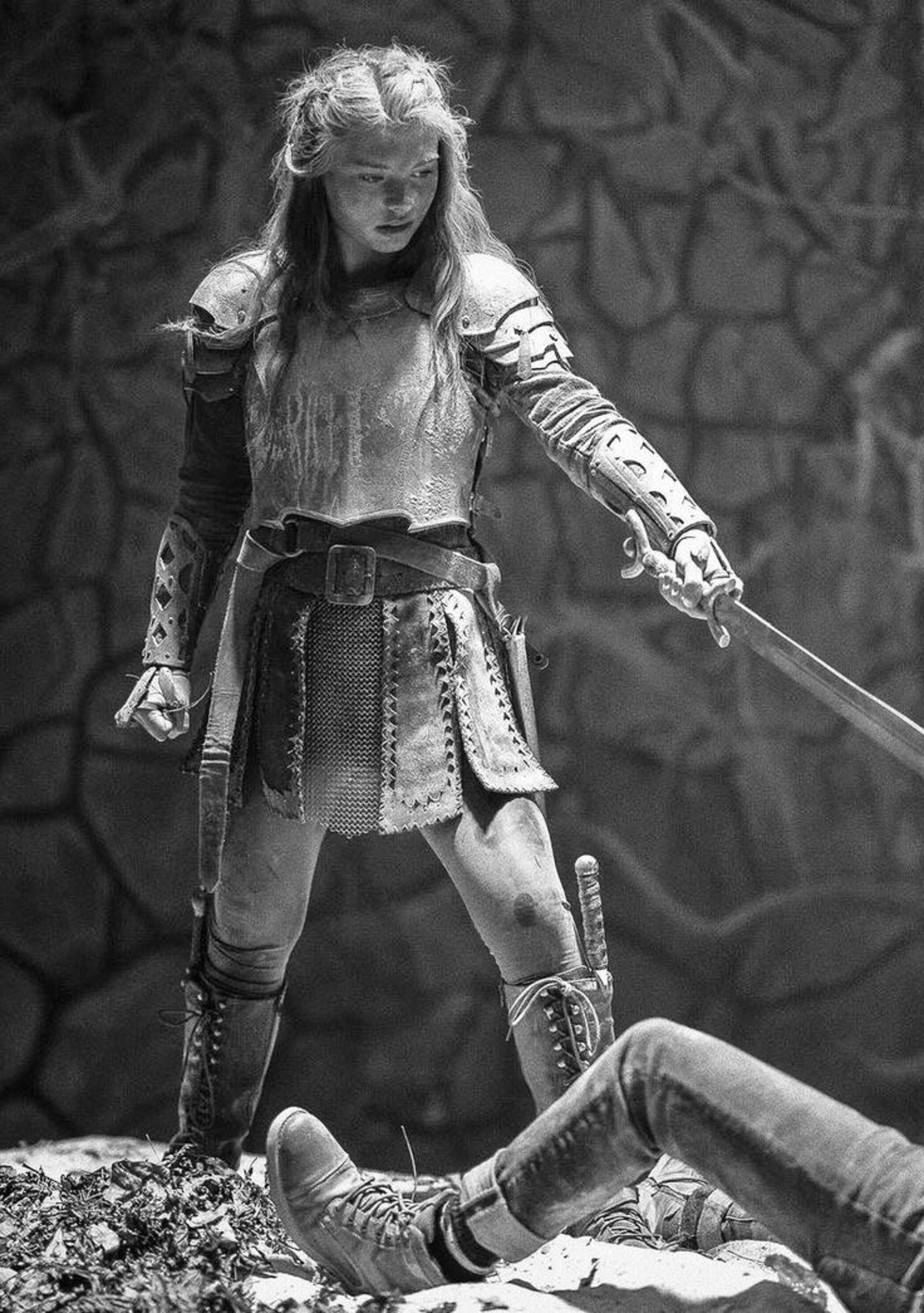 Black and white image of a young girl in armor with a sword, standing over a fallen person's legs.