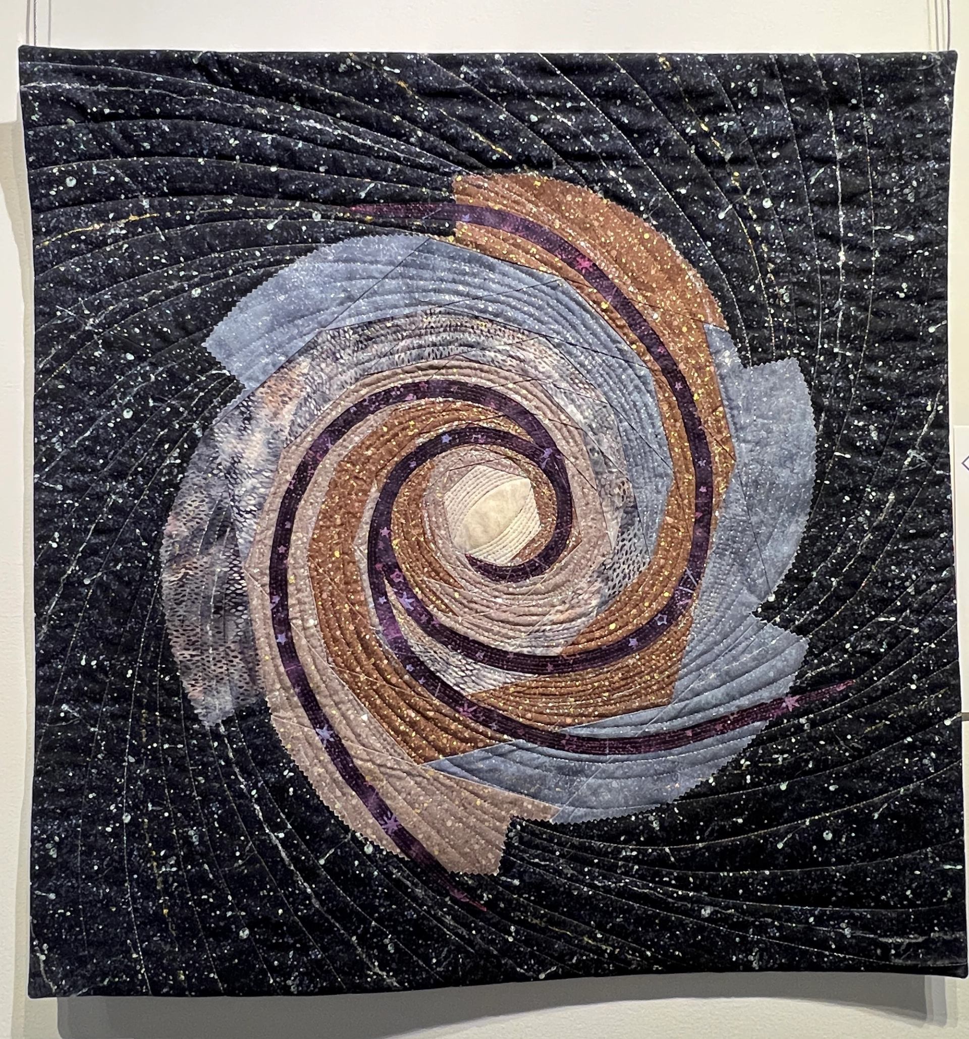 Art quilt wall hanging with concentric arcs in purple, gold, and blue on dark fabric.