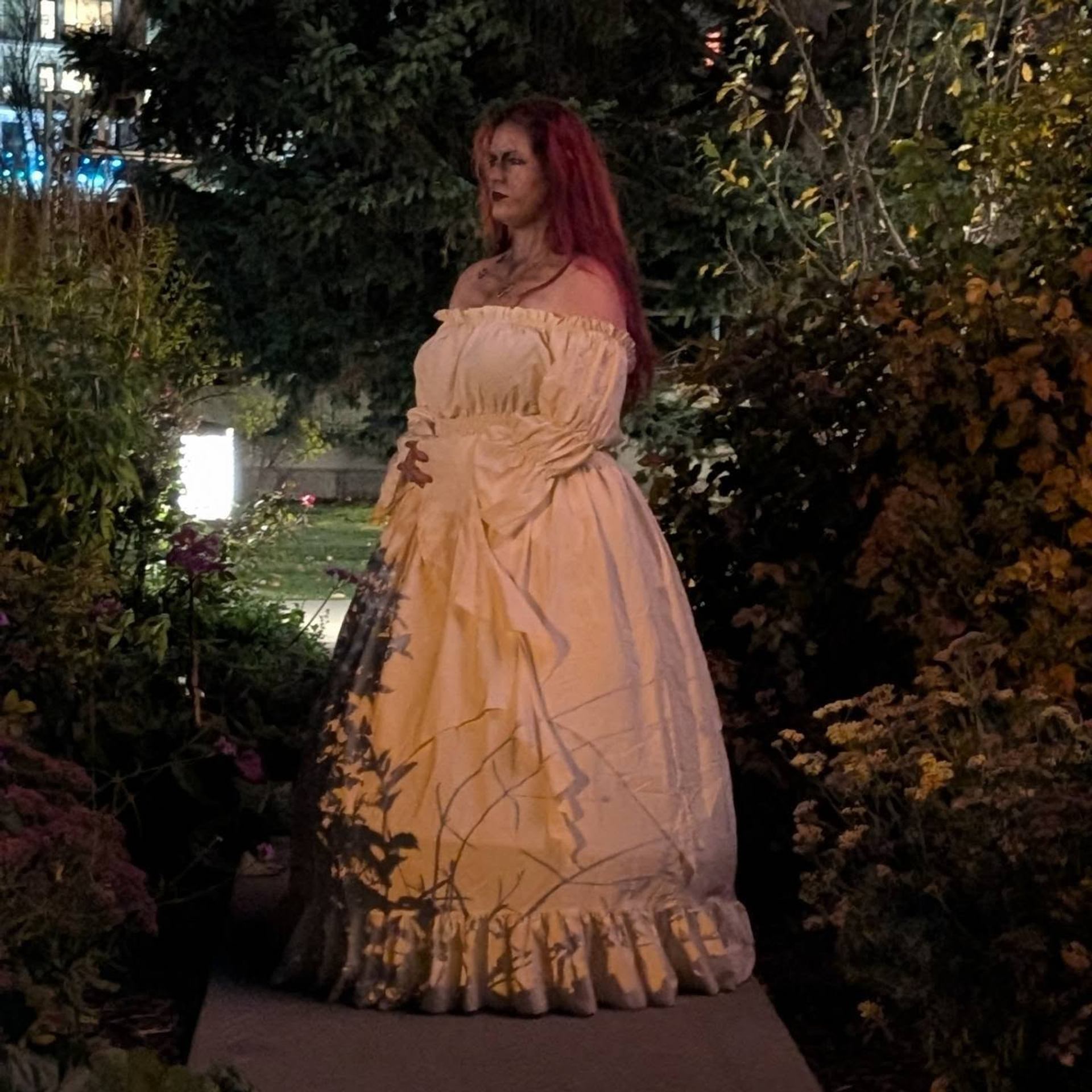 Person with long red hair wears a ruffled cream gown among dense garden foliage.