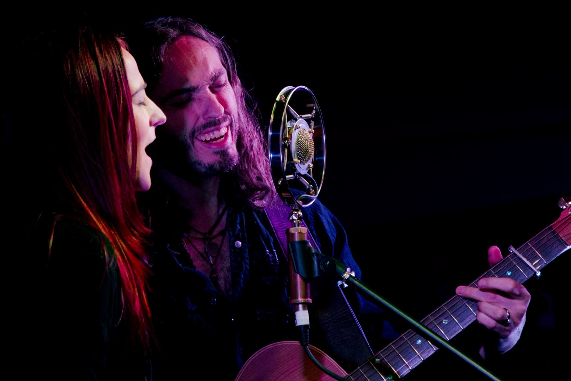 Two musicians sing into a vintage microphone while playing acoustic guitar onstage.