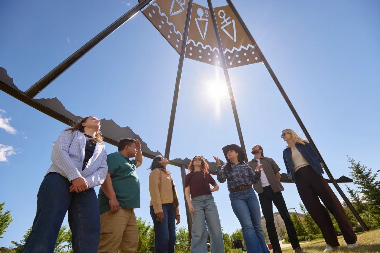 Group of people standing under large metal tipi structure with sun shining above.