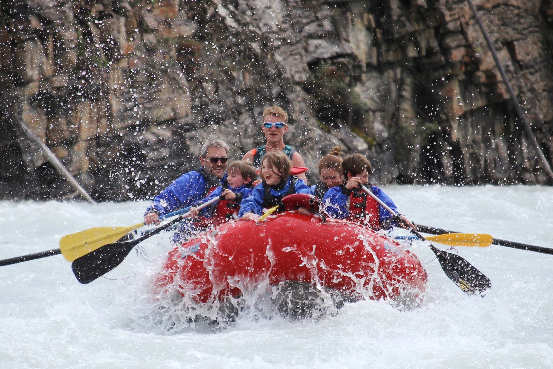 A red raft with six people, including adults and children, splashes through white water rapids.