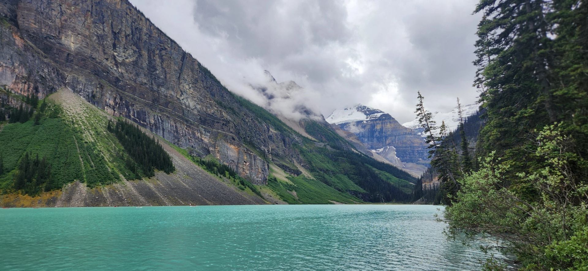 Emerald lake framed by steep rocky slopes and snow-capped peaks under cloudy skies.