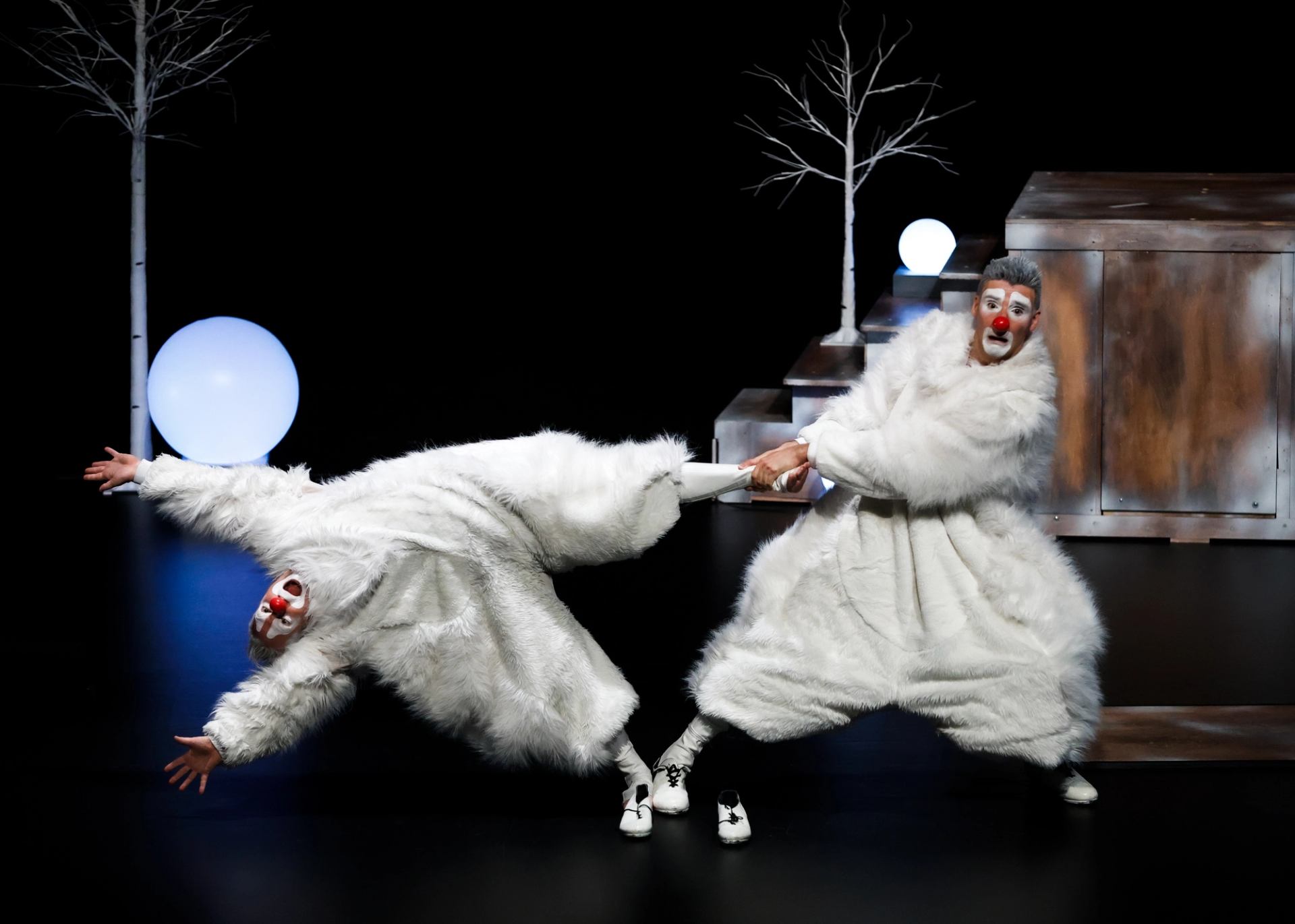 Two clowns in white furry costumes tug arms onstage beside bare trees and glowing orbs.