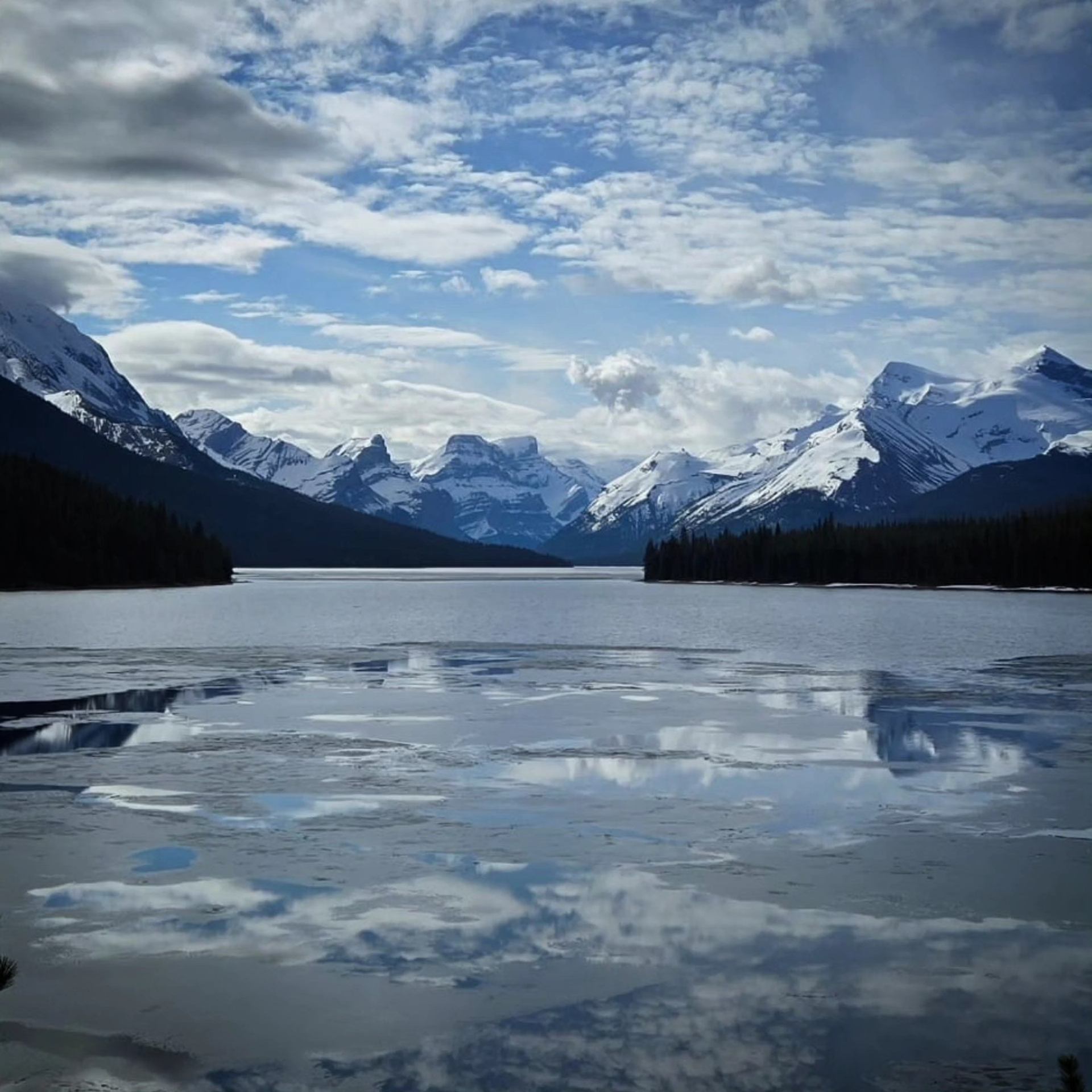 Mountain lake with reflections, pine trees, and snow-dusted peaks under cloudy blue sky.