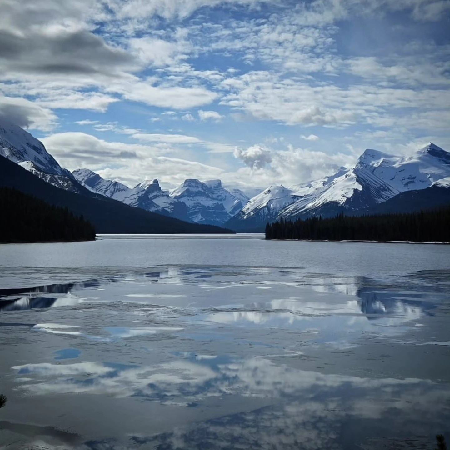 Mountain lake with reflections, pine trees, and snow-dusted peaks under cloudy blue sky.