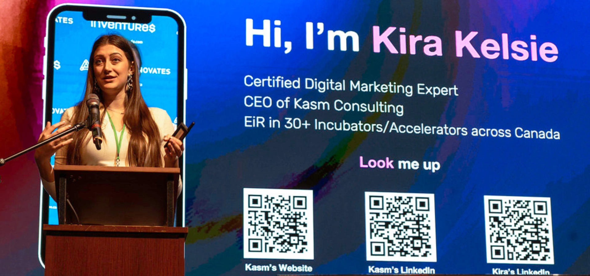 Speaker presenting at a podium in front of a large screen introducing Kira Kelsie and her roles.