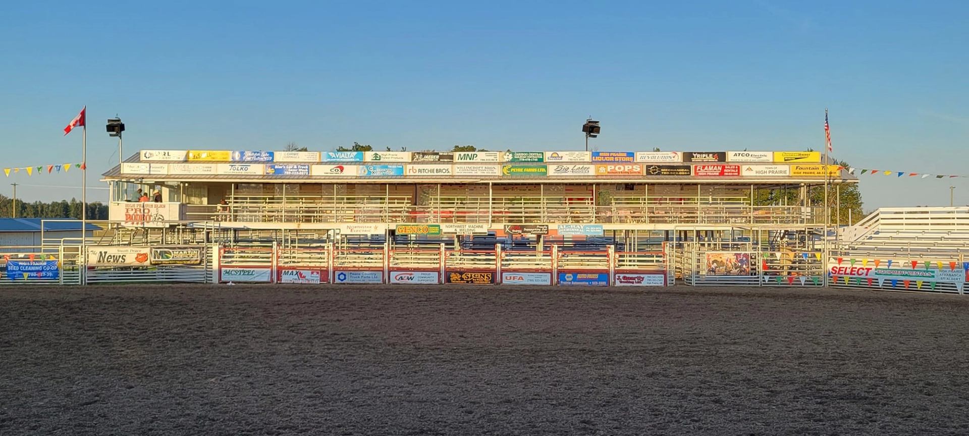 Rodeo grandstand with sponsor banners, rail fencing, and Canadian flag at High Prairie Elks arena.