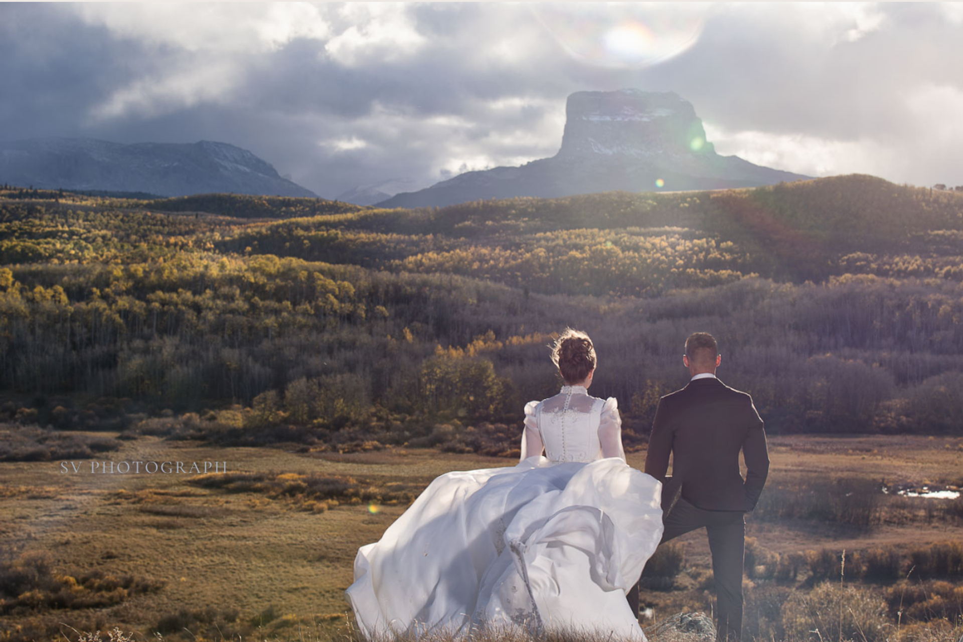 A bride and groom walk hand in hand through open land toward mountains and a glowing autumn horizon.