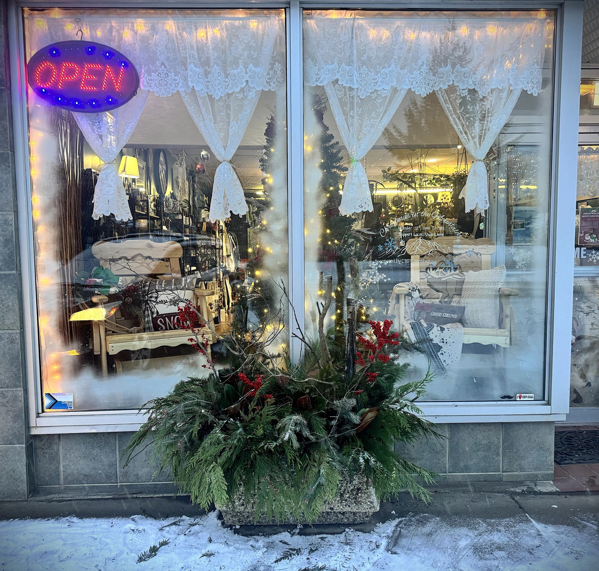 Storefront with lace curtains, glowing lights, and a winter greenery arrangement outside.