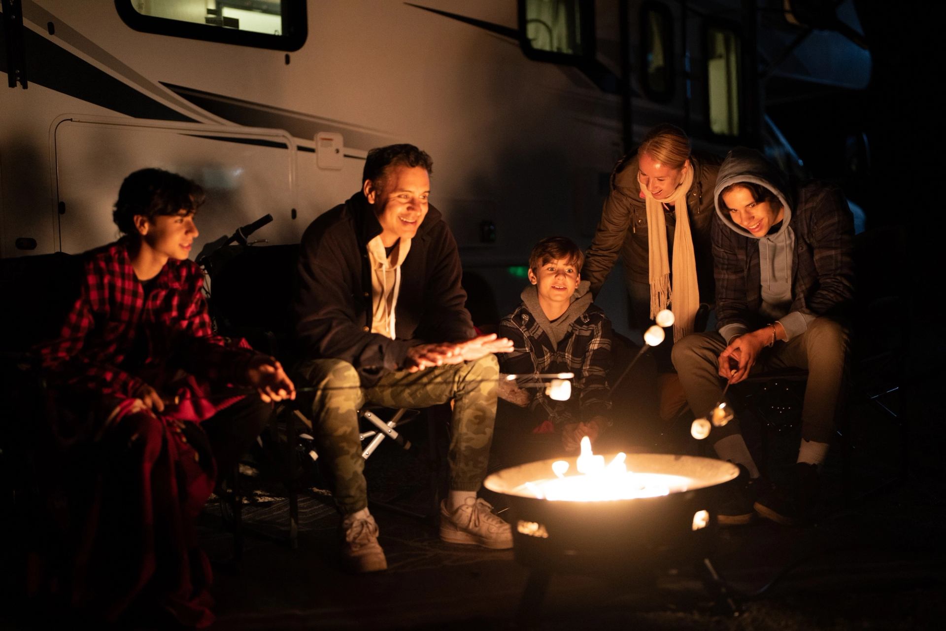 Group roasting marshmallows by a campfire in front of an RV at night.