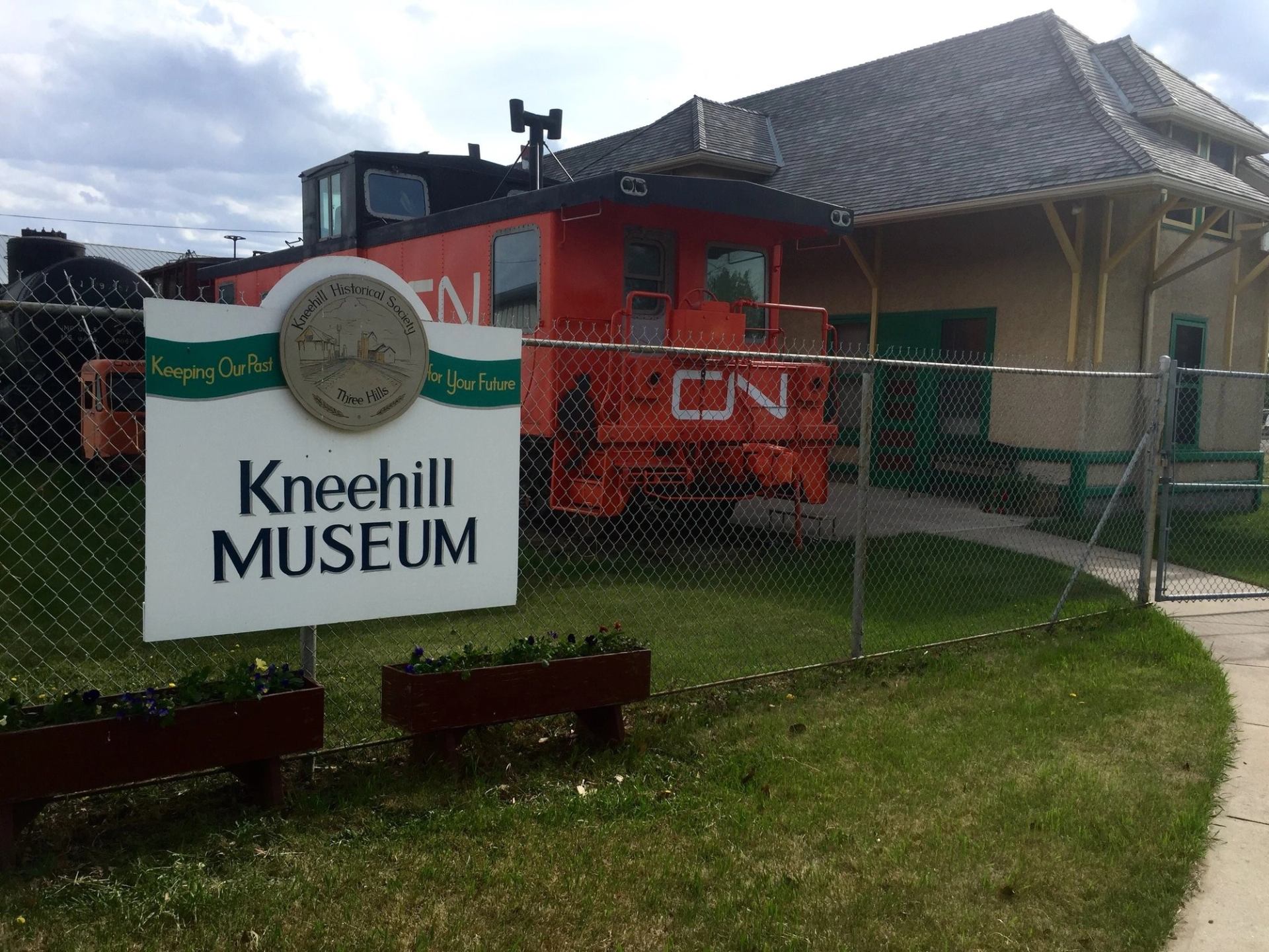 Kneehill Museum sign beside chain-link fence, red CN railcar and building.