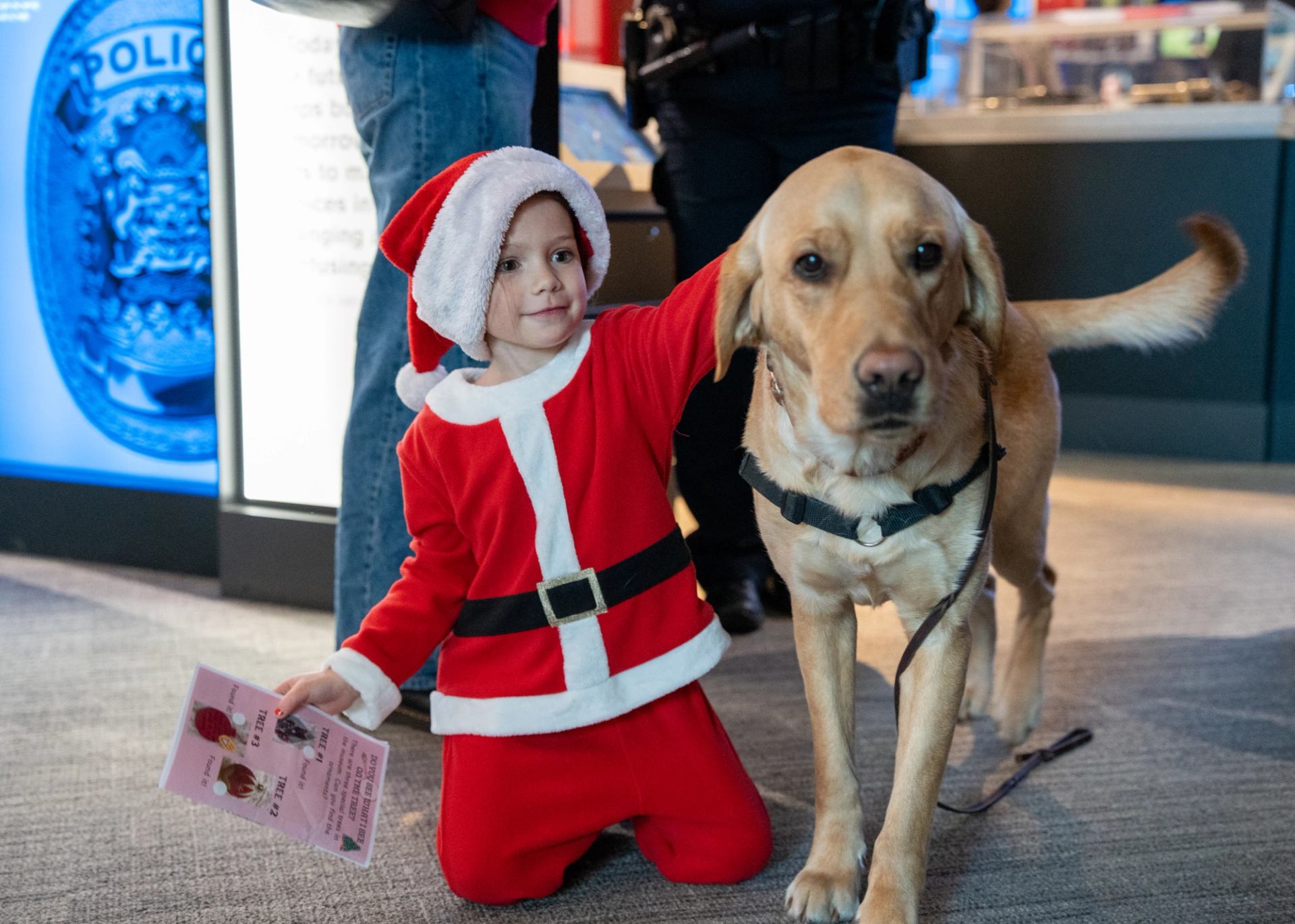 Child in a Santa costume kneels beside a yellow Labrador wearing a harness.