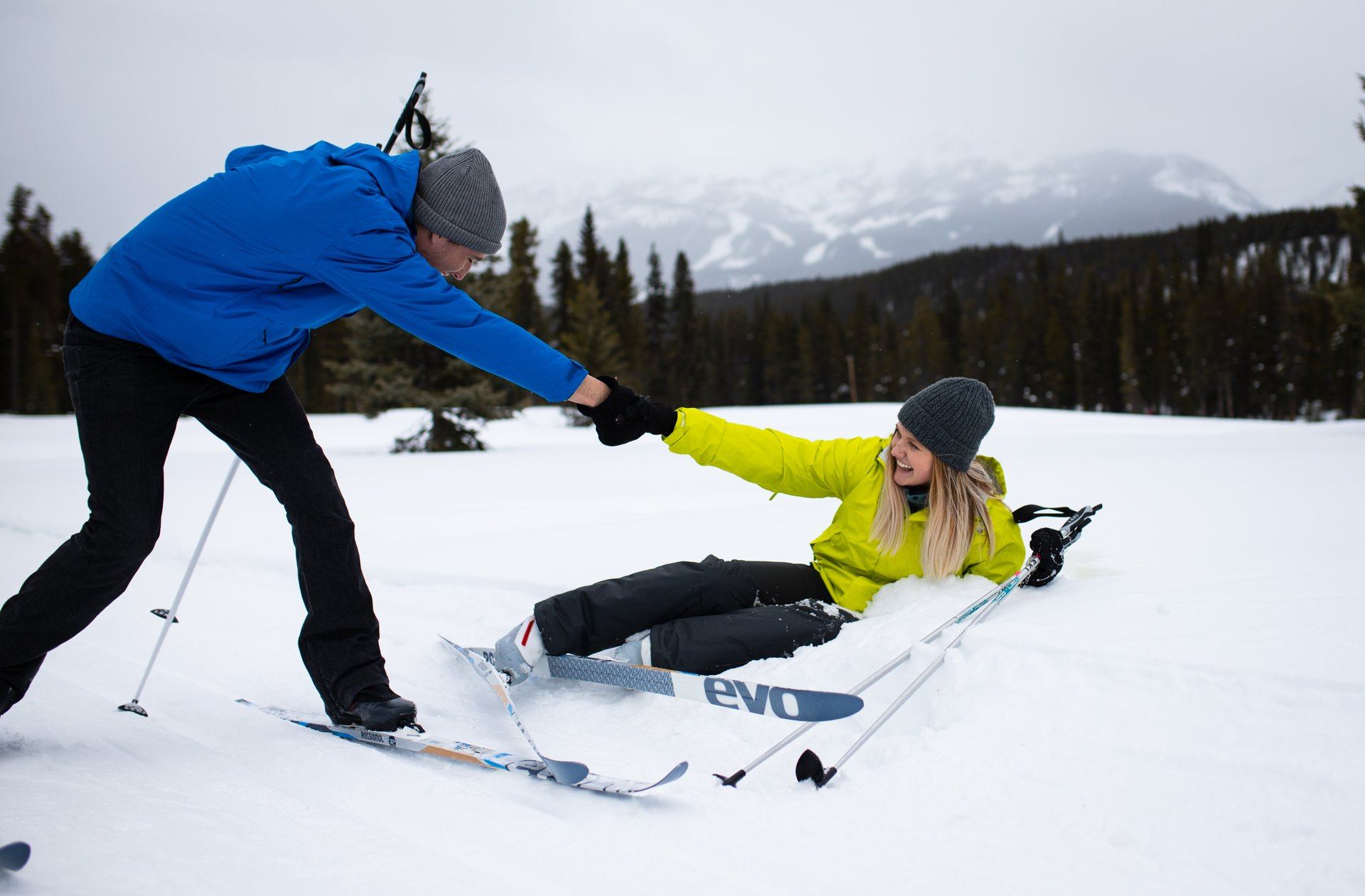 Person helping another up after a fall while skiing on a snowy trail with trees and mountains in Banff.