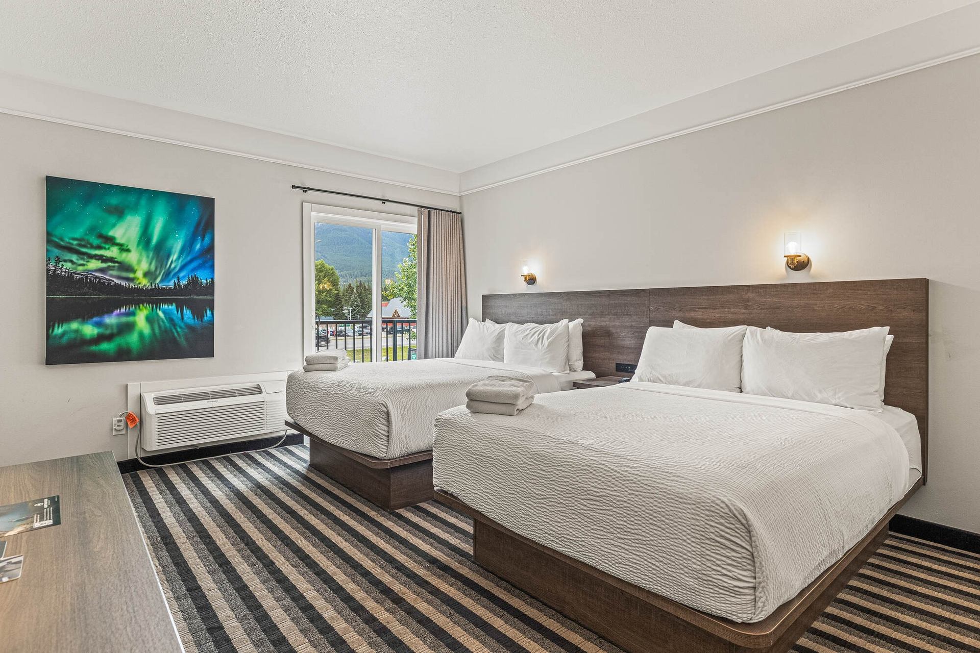 Hotel room with two double beds, striped carpet, balcony view and aurora landscape artwork.