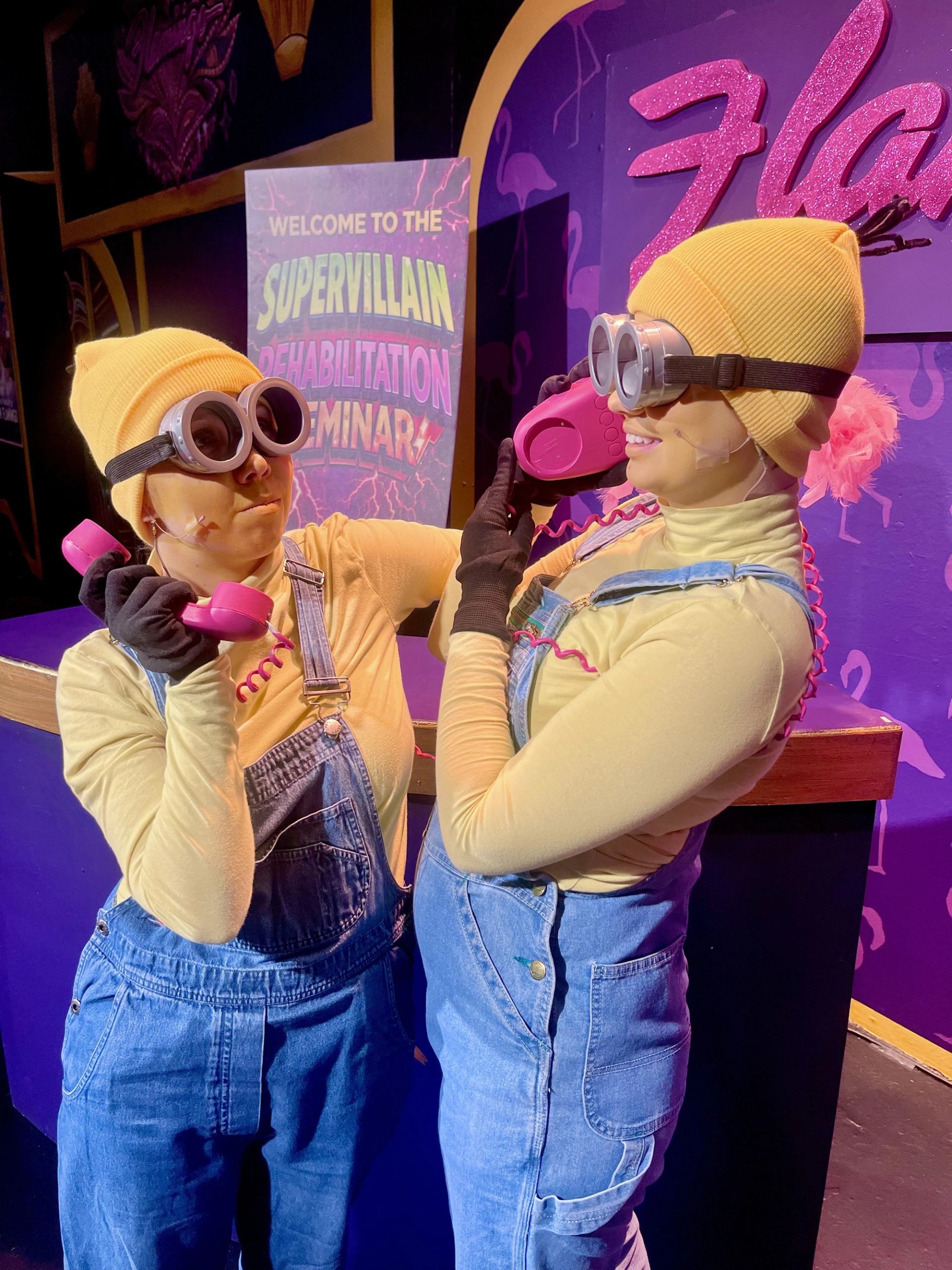 Costumed performers in goggles and overalls hold pink telephones on a colourful stage.