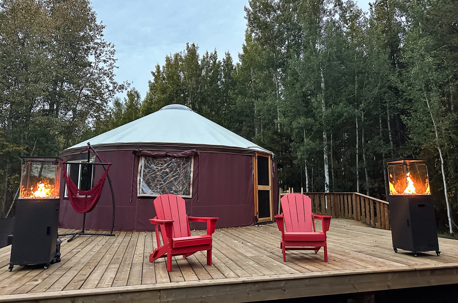 Yurt, red chairs and two fire pits sit on a wooden deck among trees.