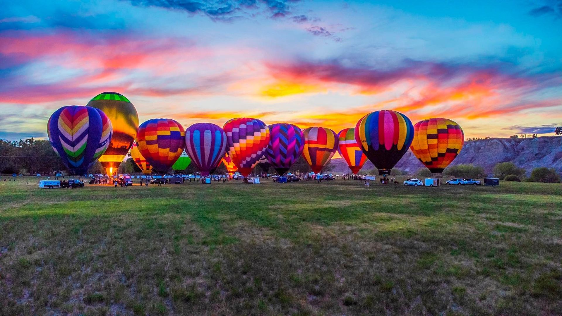Rows of colourful hot air balloons glowing at sunrise over an open field.