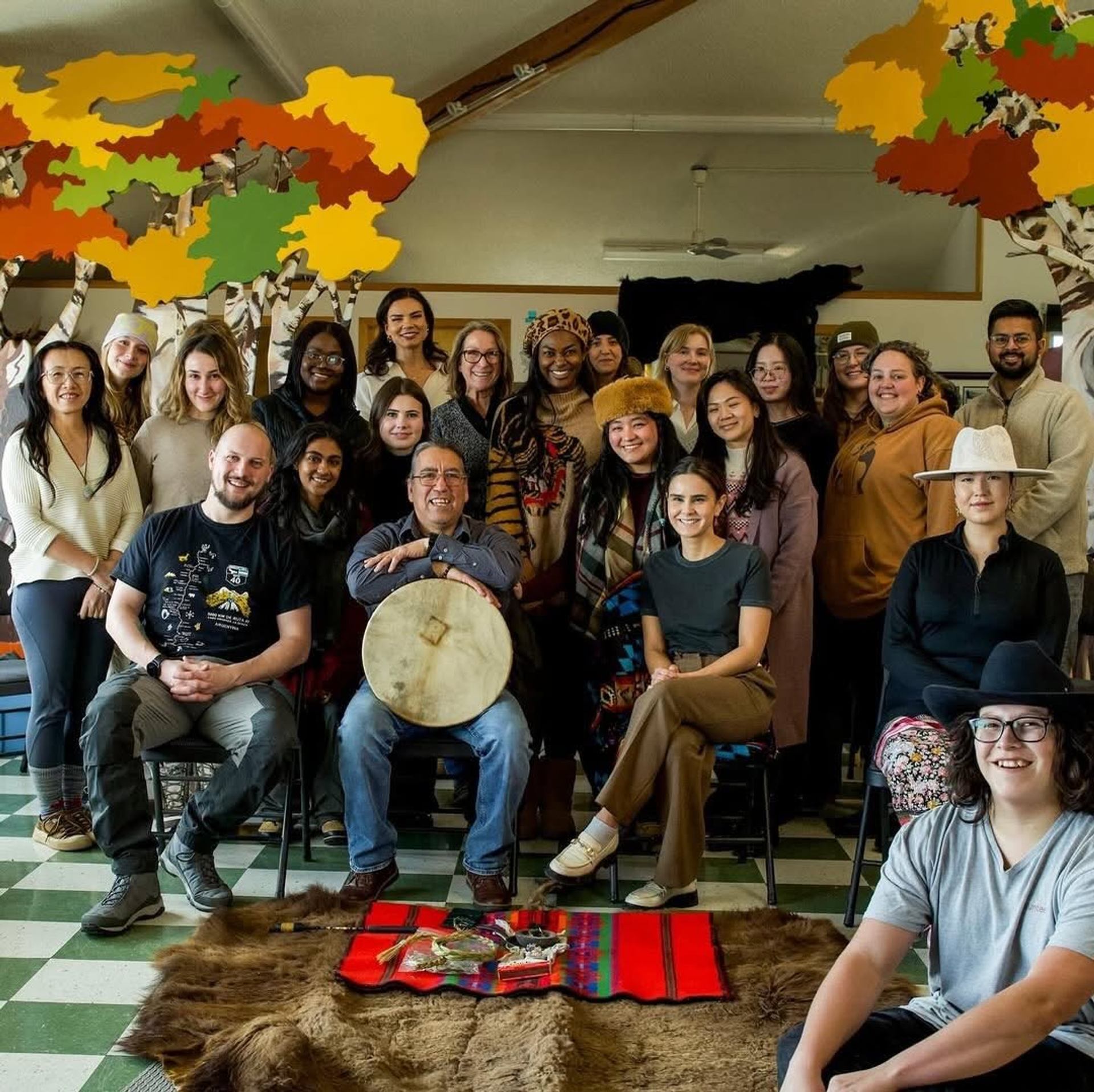 Group of people pose indoors behind drum, fur hide and colourful woven blanket.