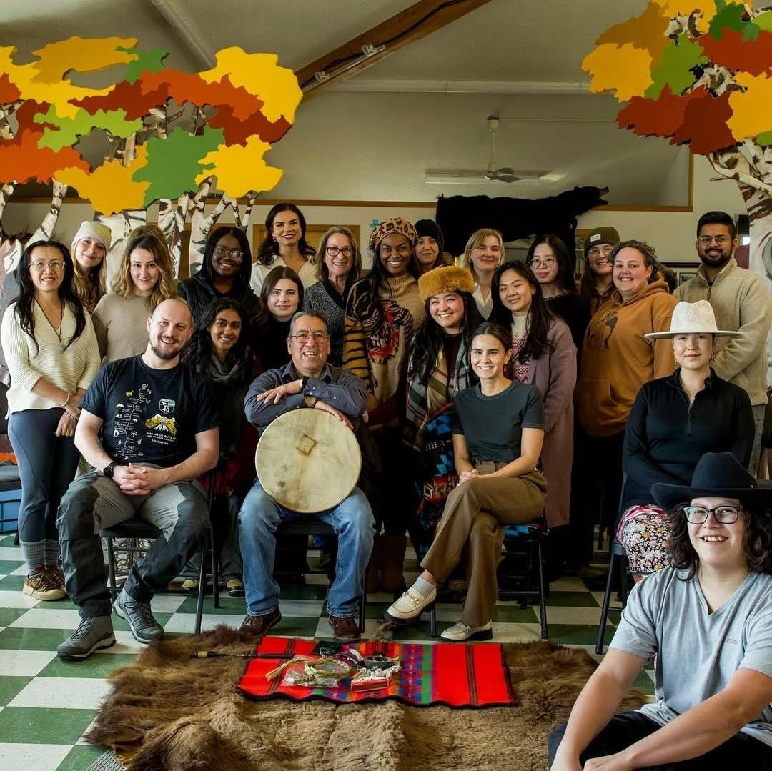 Group of people pose indoors behind drum, fur hide and colourful woven blanket.