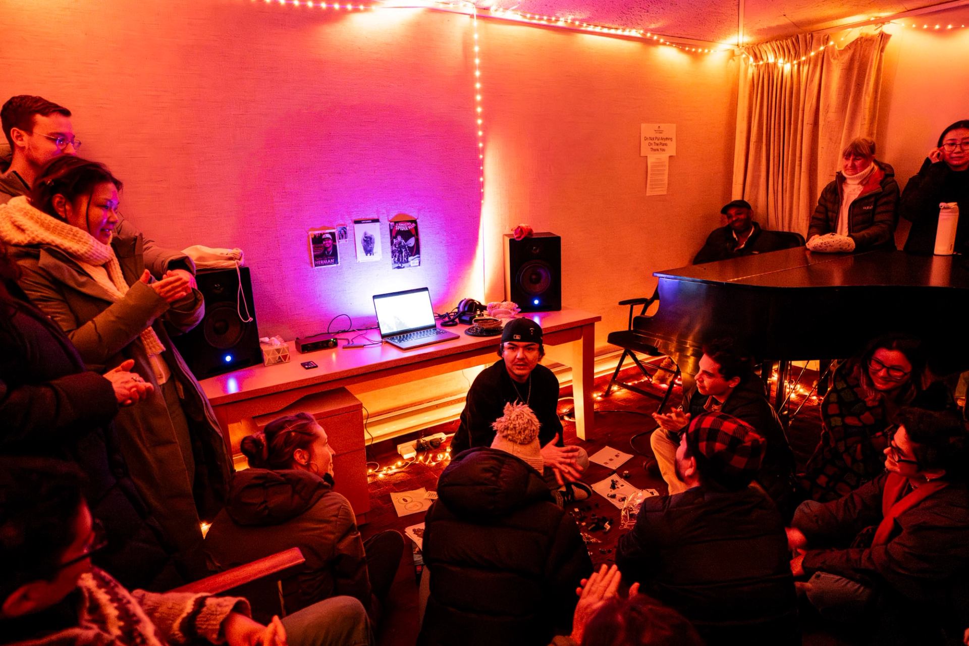 Musicians and guests gather in a warmly lit studio with a piano, speakers, and instruments during an open studio session.