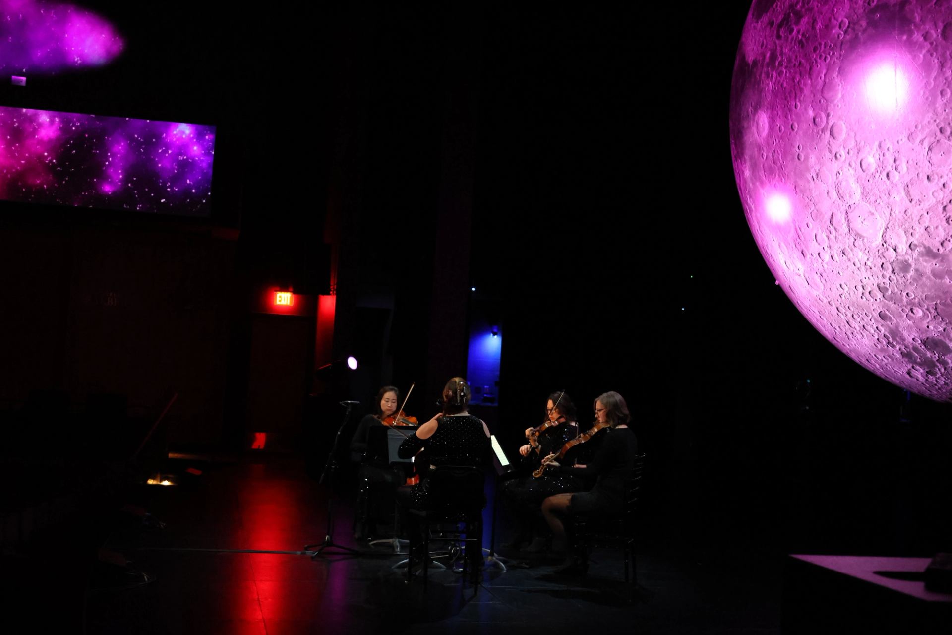 String quartet performs on dark stage beside large pink moon projection and galaxy screen.