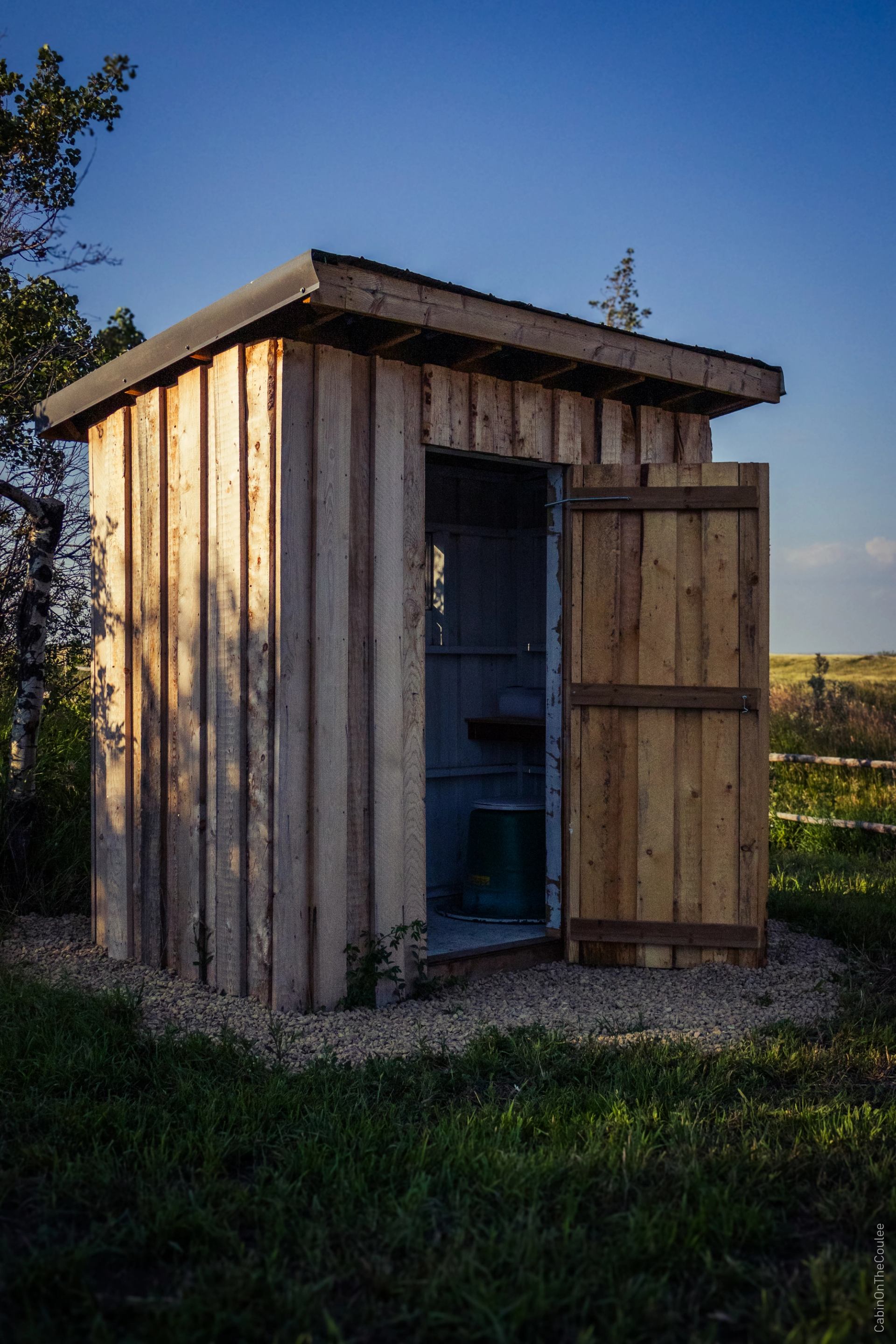 Small wooden outhouse with an open door on grassy ground at sunset.