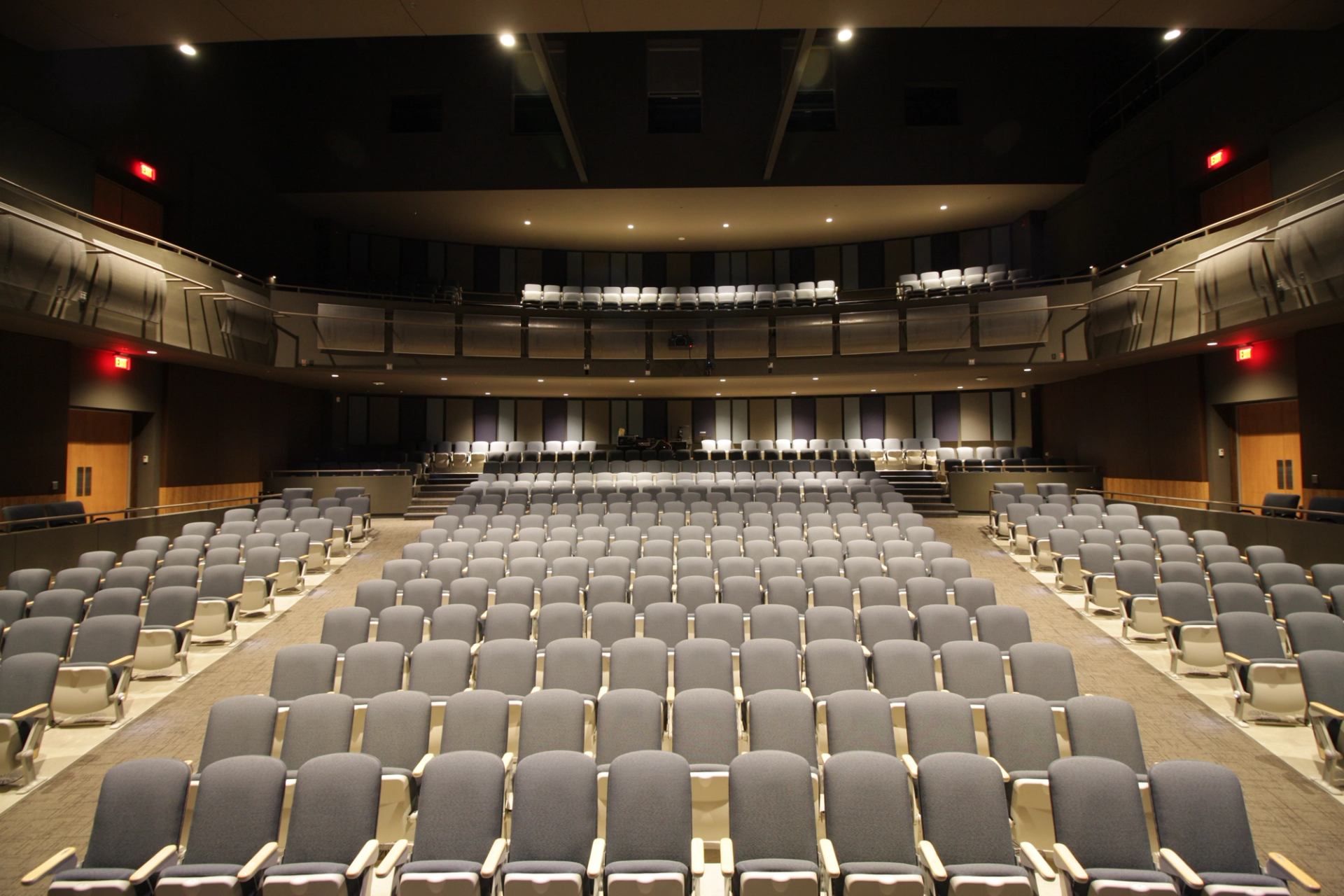 Empty auditorium with grey seats, balcony level, and illuminated exit doors.