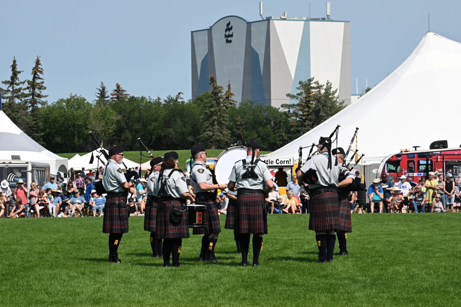 Bagpipers in kilts perform at outdoor festival with tents and crowd.