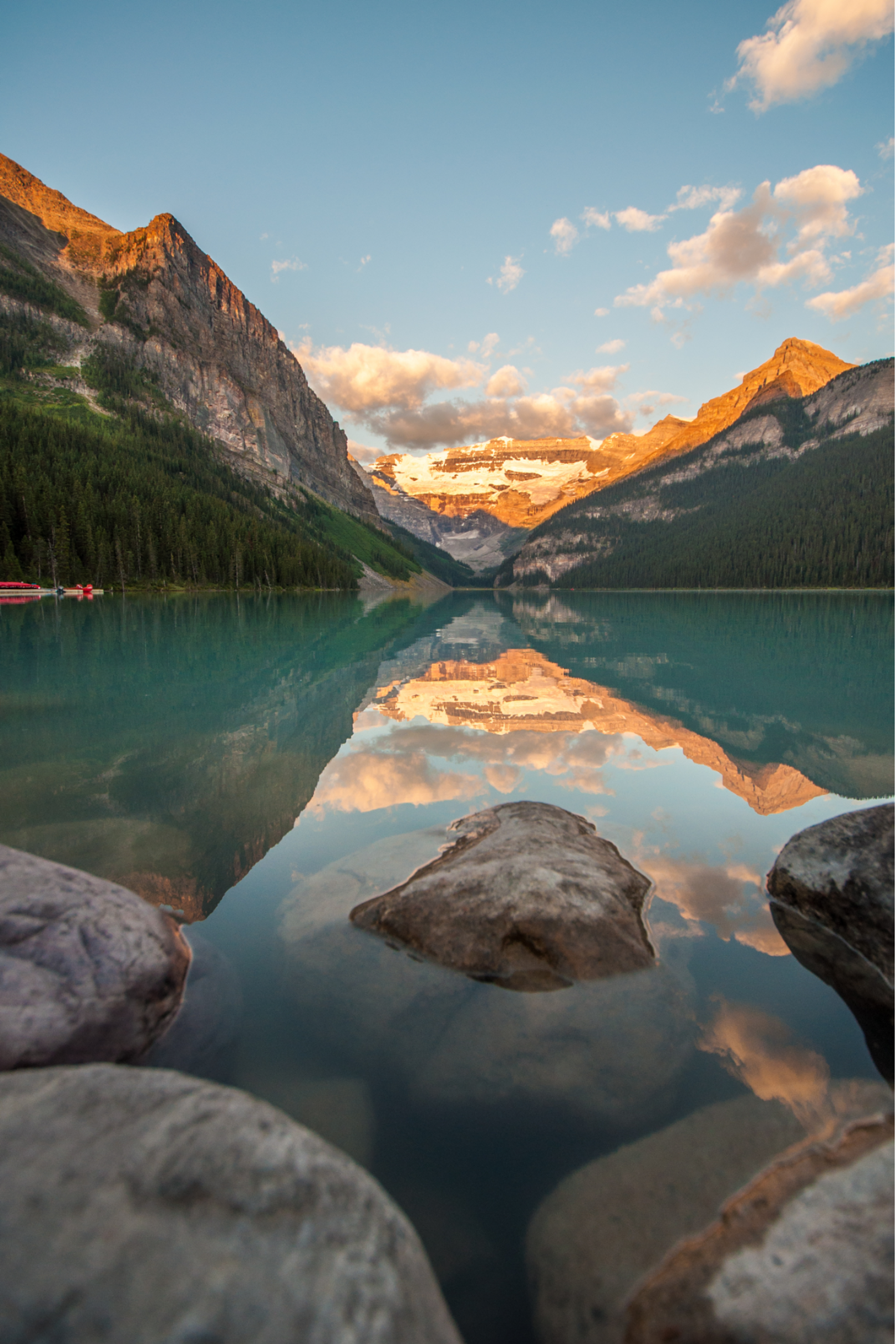 Still lake reflecting mountains and early morning light, with rocks in the foreground.