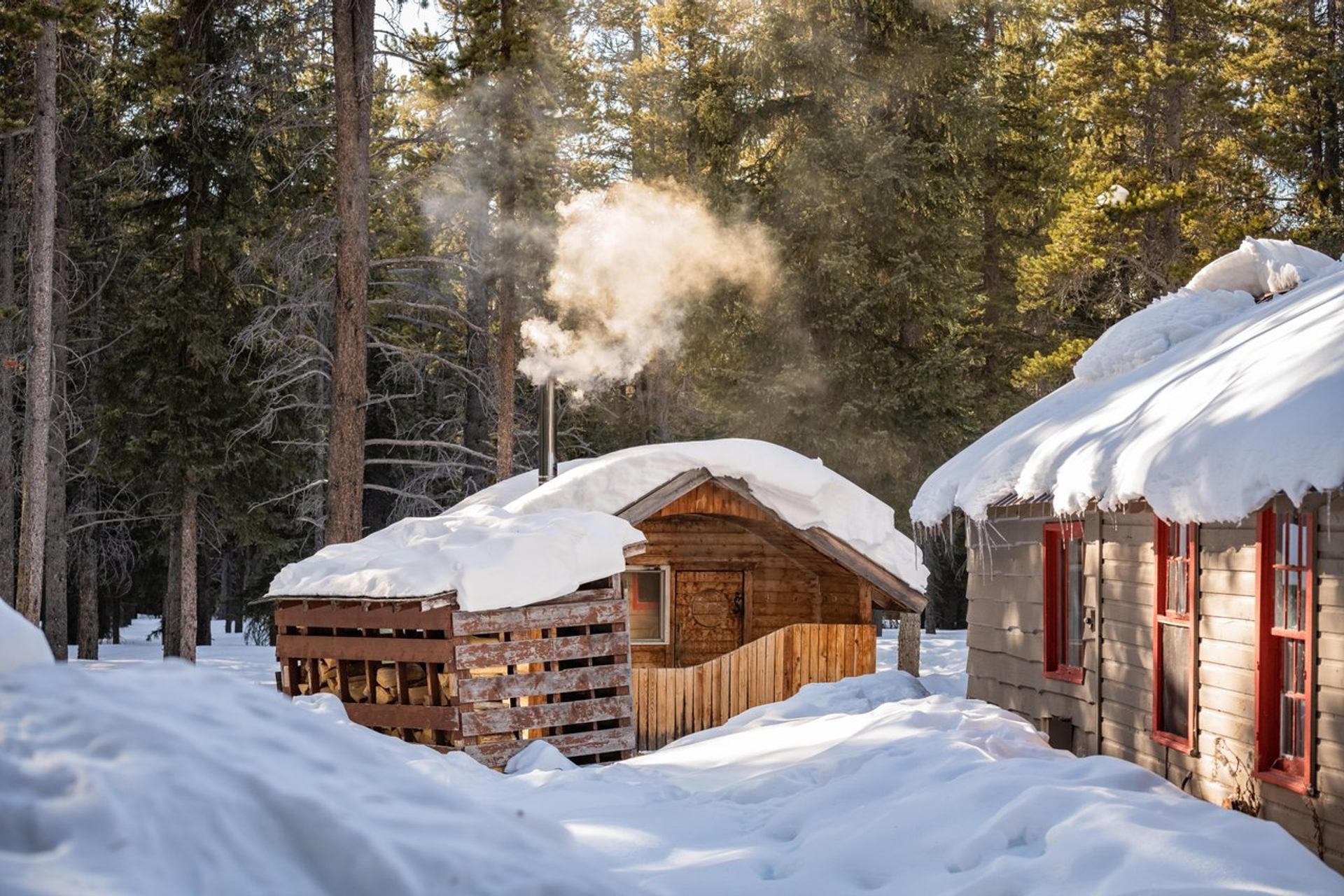 Snow-covered sauna at HI Mosquito Creek in winter with smoke rising from chimney.