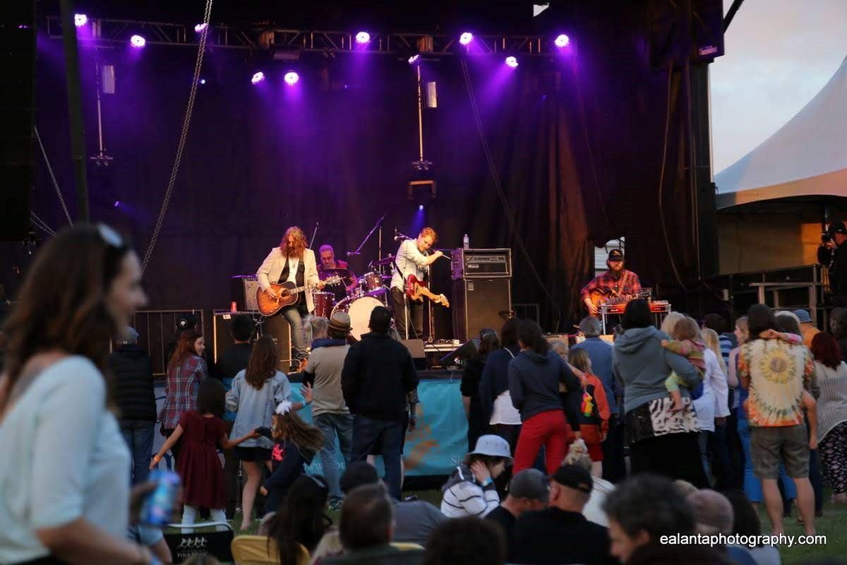 Live band performs on stage with guitar and drums at outdoor music festival, audience seated and sta.