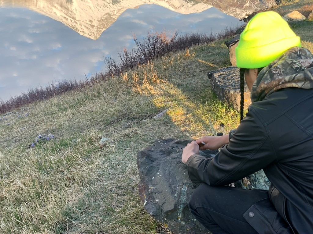 Person in outdoor gear sitting on a rock with a bright neon hat, overlooking grassy terrain and mountains.
