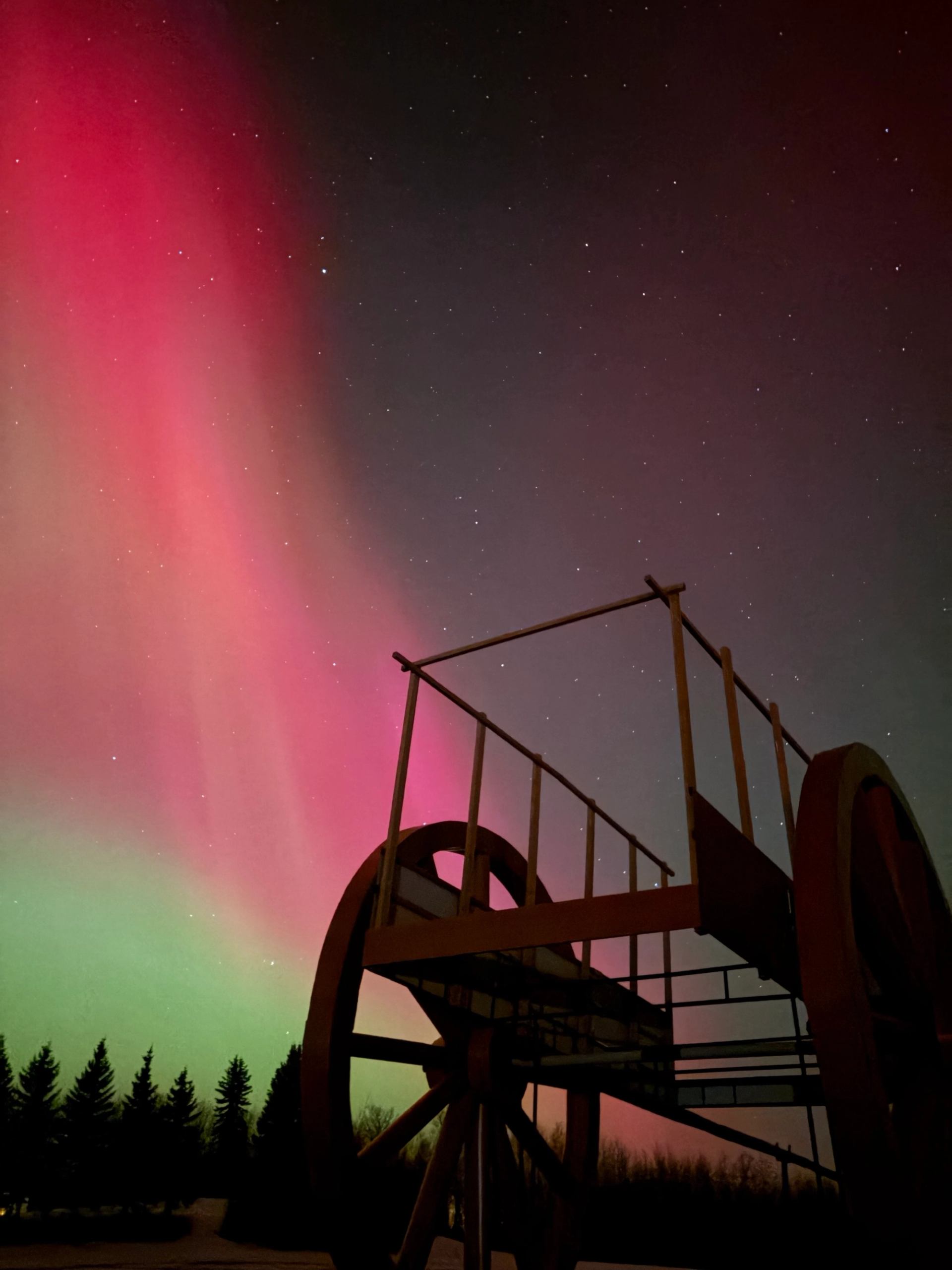Silhouette of Alberta’s Largest Red River Cart beneath vivid red and green northern lights.